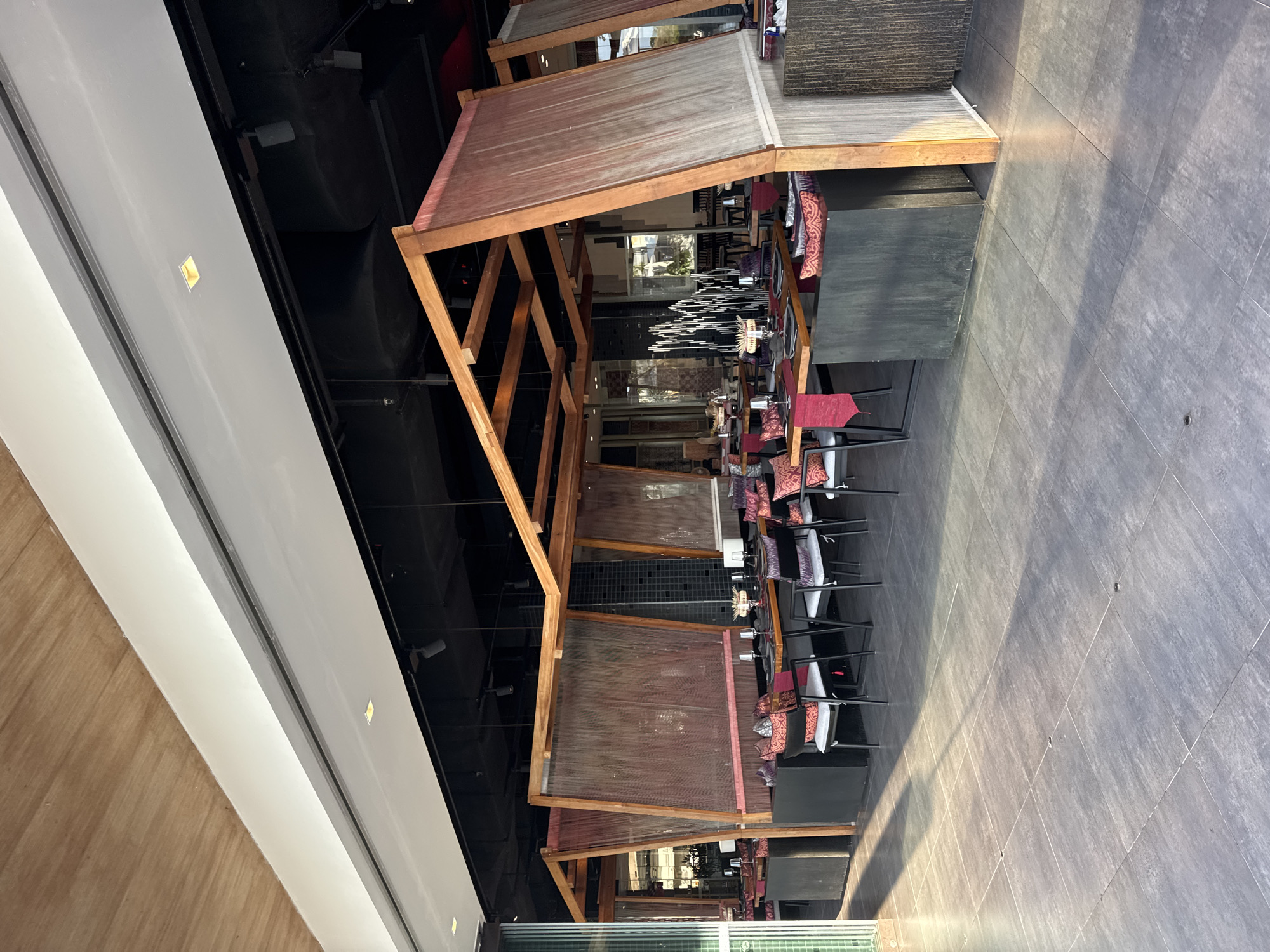 The image shows an outdoor seating area of a restaurant in Kuta Selatan, Indonesia. The area is furnished with several dining tables and chairs, all set for meals. The tables are adorned with red placemats, napkins, and decorative pillows, suggesting an elegant dining experience. The seating area is partially covered by wooden structures and sheer fabric curtains, creating a semi-private ambiance. In the background, a water feature adds a touch of serenity to the scene. The floor is paved with large gray tiles, and sunlight casts long shadows across the area, indicating it is either morning or late afternoon. The restaurant appears to be empty, with no customers or staff visible.