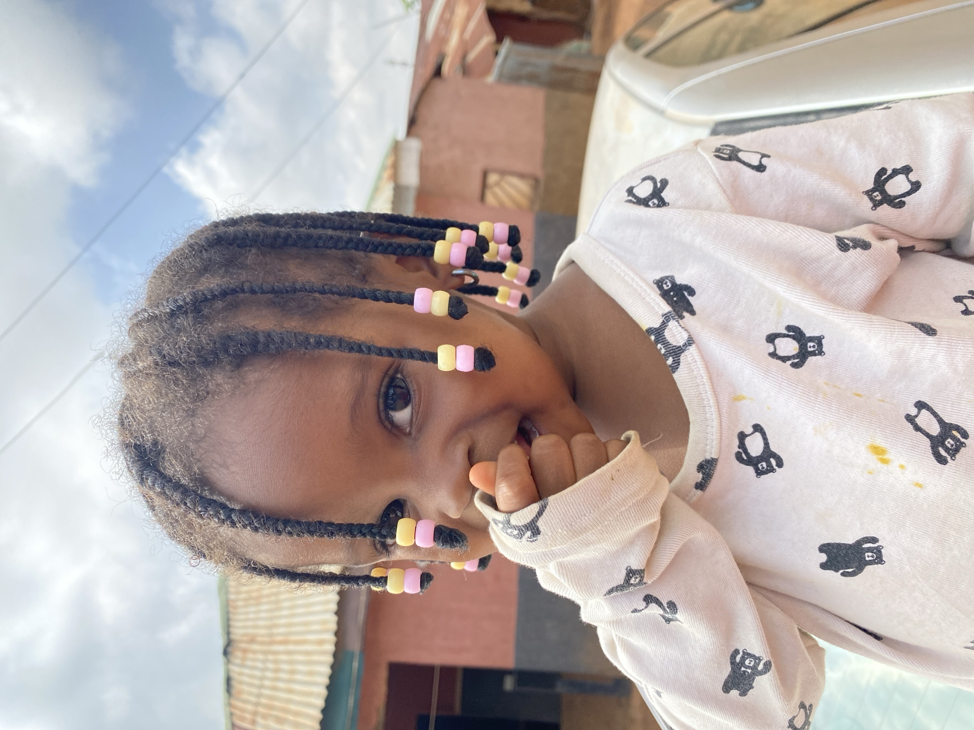 The image shows a close-up, rotated view of a young Black girl with braided hair adorned with pink and yellow beads. She is wearing a light-colored long-sleeved shirt with a repeating pattern of black bear figures. Her hand is near her mouth, suggesting she might be playing or eating. The background is blurred but shows hints of an outdoor environment with a corrugated metal roof and a pink building, consistent with the Bukuru, Nigeria location context. The sky with clouds is visible above, indicating it is daytime. The overall mood appears playful and candid. There is no visible text in the image.