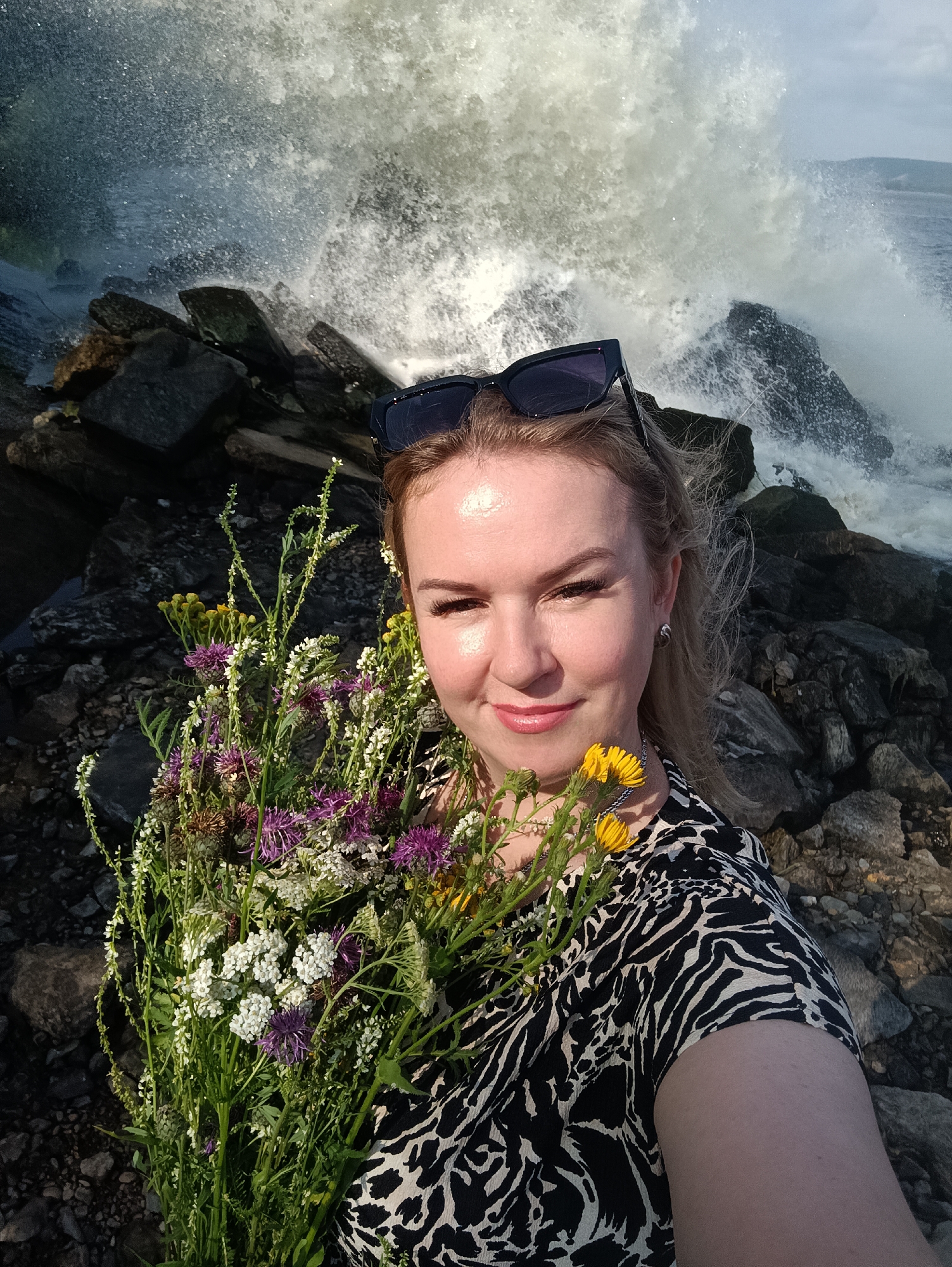 A blonde woman in a black and white patterned dress poses for a selfie while holding a bouquet of wildflowers. She is wearing black sunglasses on her head and a silver earring. She is standing on a rocky shore with a large wave crashing behind her. The location is the city of Chernushka, Russia.