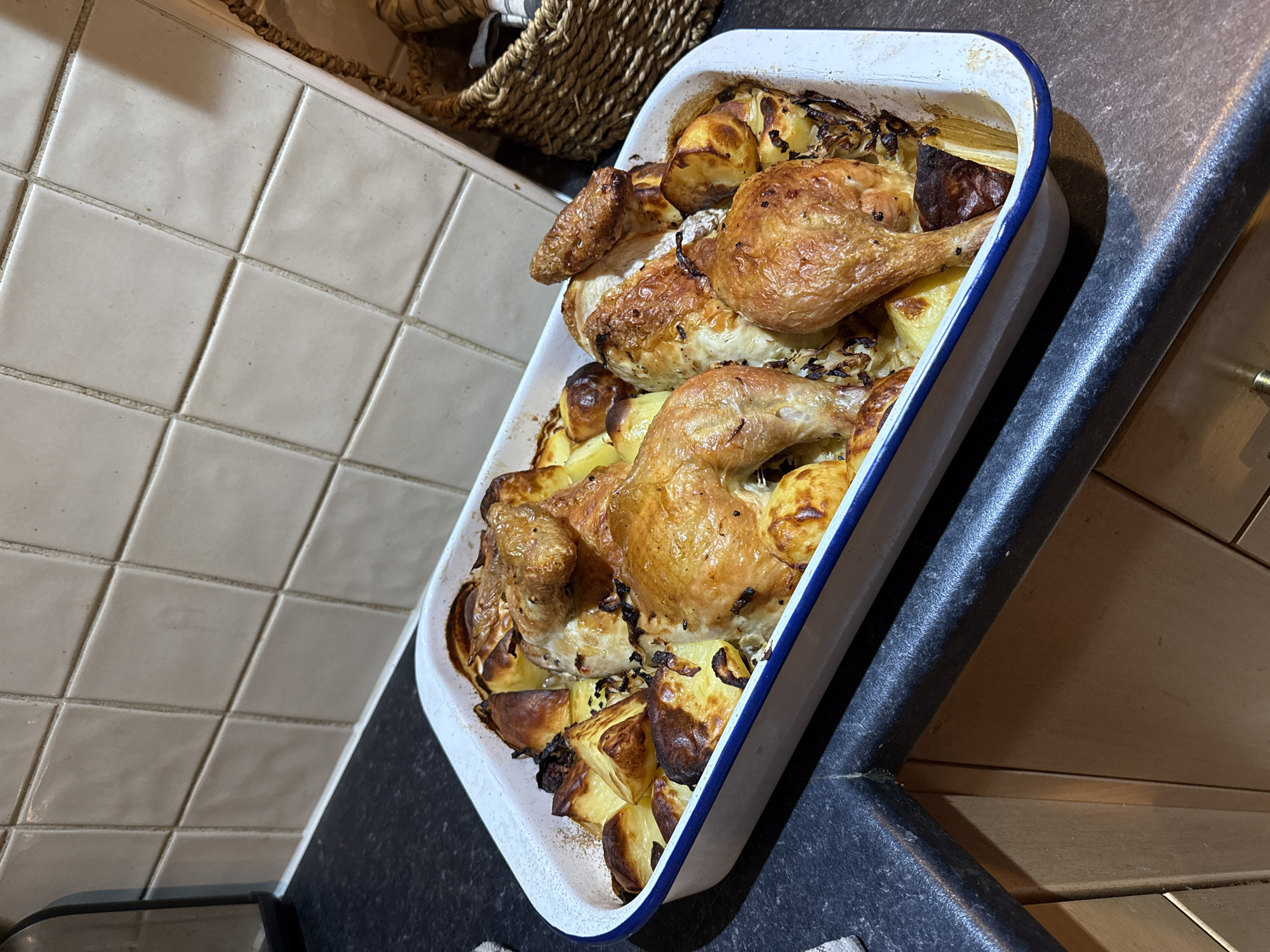 A close-up, slightly angled shot shows a white rectangular baking dish with a blue rim filled with roasted chicken pieces and potatoes. The chicken has a golden-brown, crisp skin. The potatoes appear to be roasted to varying degrees of doneness, some with dark, crispy edges. The dish is placed on a dark countertop or surface. In the background, there is a tiled wall to the left and a woven basket. No people are visible. The environment suggests a domestic kitchen setting. The location in the city of Great Yarmouth, United Kingdom, is not discernable from the image alone.