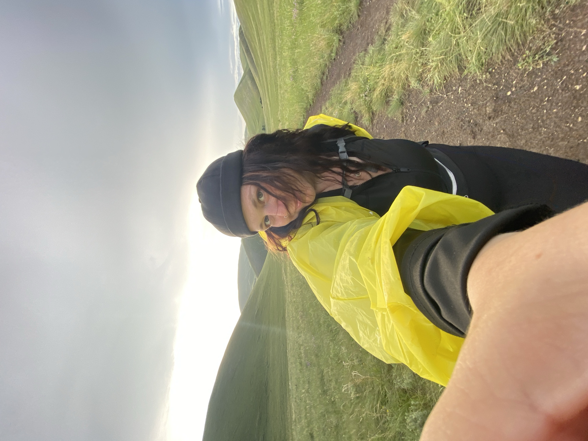 The image shows a young woman wearing a black beanie and a bright yellow hooded jacket over a black zippered vest and black leggings. She has long, dark hair and is wearing a nose ring. She is holding a camera in her left hand, extending it towards the viewer as if taking a selfie. The woman is standing on a dirt path in a vast, grassy landscape with rolling green hills in the background under a cloudy sky. The location is described as the city of Donskoe, Russia.