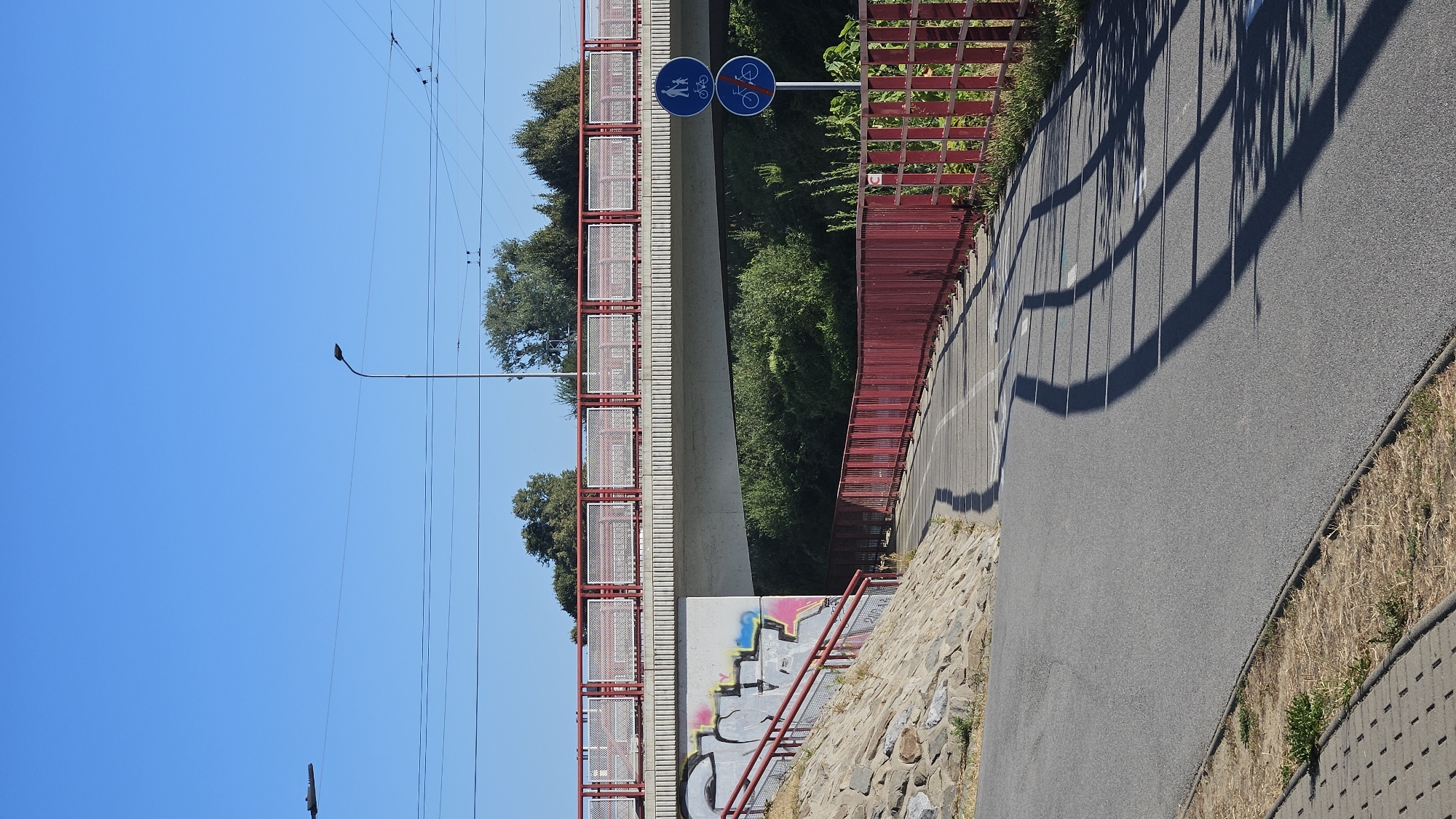 The image shows a street or pathway in Prešov, Slovakia. A concrete bridge spans the scene, supported by a concrete pillar with graffiti on its side. A red railing runs along the side of the bridge and down a ramp to the left. To the right of the bridge pillar, a pathway curves away, bordered by a red fence. Two circular blue signs are mounted on a pole next to the pathway; the top sign depicts a pedestrian and a bicycle, while the bottom sign shows a bicycle with a red line through it, indicating no cycling. The pathway appears to be a dedicated cycle path. The ground is paved with asphalt and there is dry grass and some vegetation. The sky is clear and blue, and the scene is illuminated by sunlight, casting shadows from the fence onto the pathway.