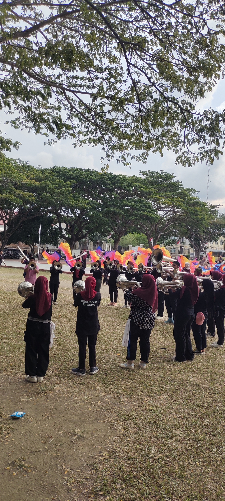 The image depicts a marching band practice or performance in an outdoor grassy field, likely in Lhokseumawe, Indonesia, given the context of the provided information. In the foreground, several individuals wearing black uniforms and headscarves are holding brass instruments, such as tubas and trumpets. One individual in the middle foreground wears a shirt with text that reads "PECOK ELORAN HITAM MARCHING BAND". In the background, more band members are visible, along with colorful flags being waved. Large trees with dense green foliage form a backdrop to the scene, and a cloudy sky is visible above. There are buildings and possibly other trees in the far background, suggesting an urban or semi-urban setting. The ground is covered with dry grass and some patches of dirt.