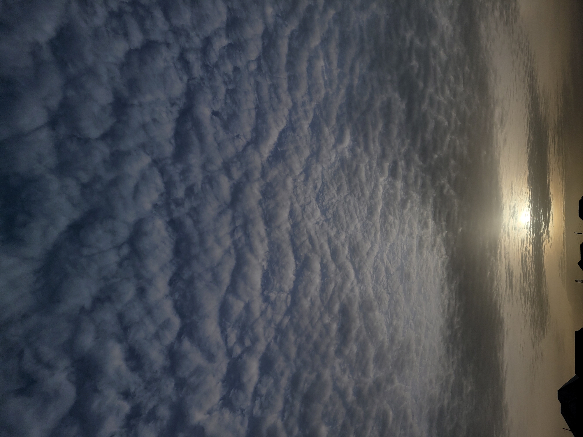 The image captures a dramatic sky filled with clouds, likely at sunrise or sunset, over what appears to be a distant horizon with structures. The sky is dominated by a vast expanse of clouds exhibiting a distinct, rippled or cellular pattern, often characteristic of altocumulus or stratocumulus formations. These clouds range in color from a deep, dusky blue-grey in the upper left to brighter, luminous white and grey tones closer to the sun.
The sun is positioned towards the right side of the frame, partially obscured by the clouds, but its intense light shines through, creating a brilliant, hazy glow. This illumination gives a warm, golden cast to the clouds and atmosphere around it. Along the very bottom edge of the frame, dark silhouettes of various structures, possibly buildings, rooftops, or communication towers, are visible against the brighter sky, hinting at a populated area. No specific activity or event beyond the natural celestial display is depicted, and no people are visible. The scene conveys a sense of atmospheric beauty and grandeur. There is no visible text in the image. Given the provided context, this atmospheric scene is located in Barakin Akawo, Nigeria.