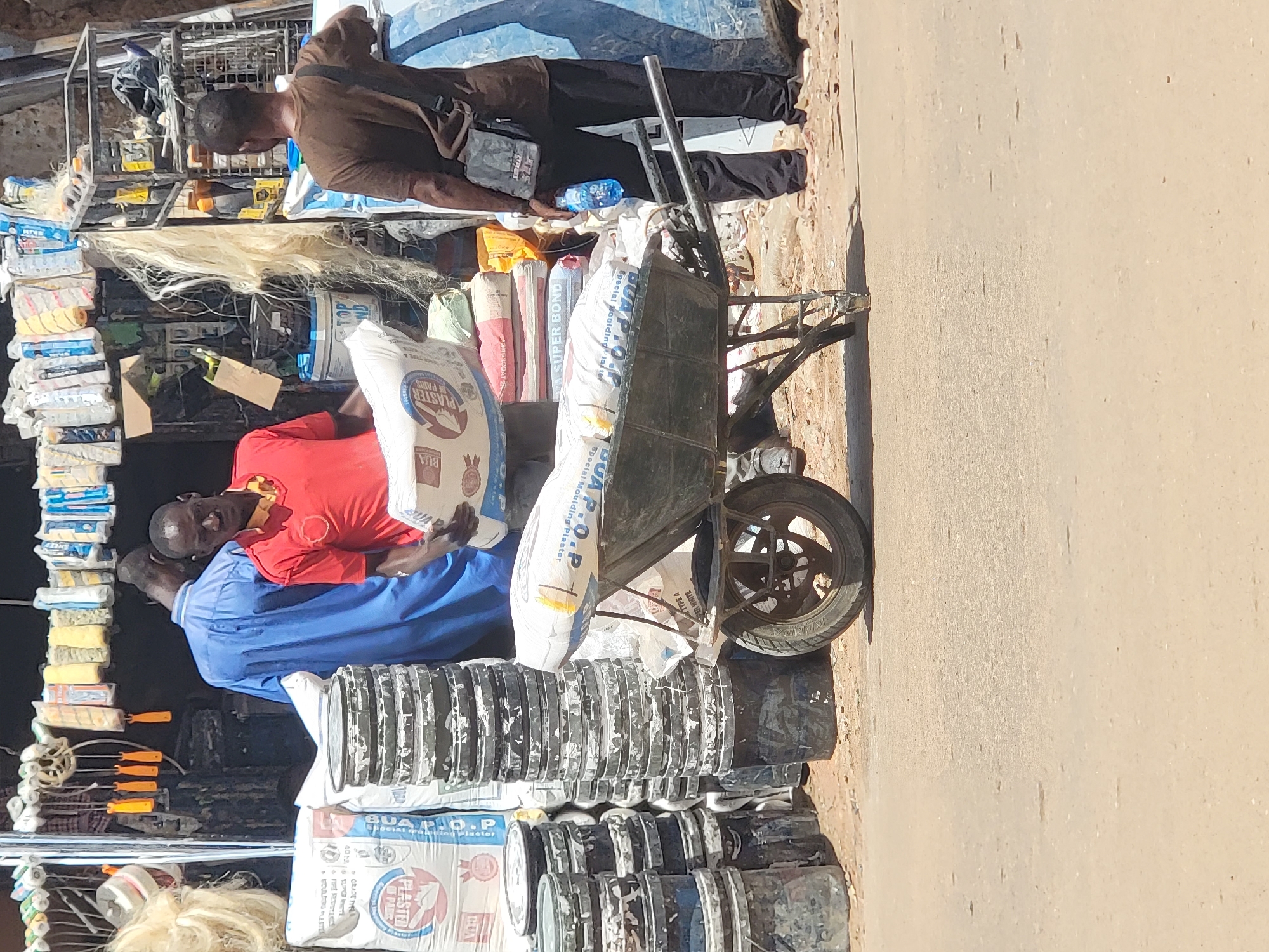 A street vendor in Maiburiji, Nigeria, is selling construction materials. One man in a brown shirt is looking at the merchandise. Another man wearing a red shirt over a blue shirt is holding a bag of "PLASTER of Paris." A wheelbarrow loaded with bags of "BUA P.O.P Special Moulding Plaster" and other materials sits on the side of a dusty road. There are cans and stacks of supplies on display.