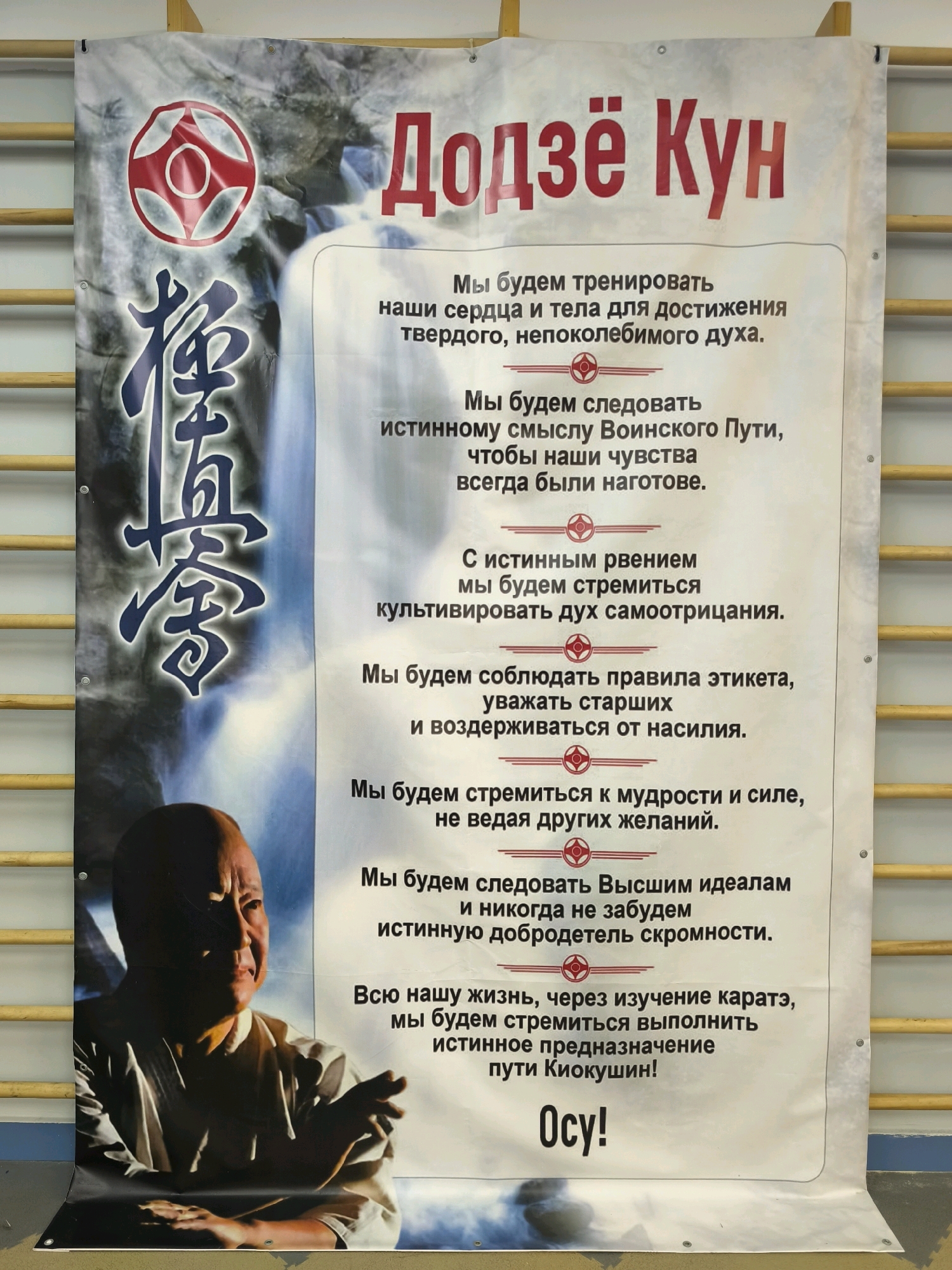 The image shows a banner with Russian text and graphics, likely for a martial arts dojo. The banner features a large Japanese kanji character on the left, a red and white circular emblem above it, and a black and white portrait of an Asian man in the lower left corner. The right side of the banner contains several lines of text written in Russian. The banner is hung on a wall with wooden bars. The background appears to be a depiction of a waterfall. There is no indication of the location being Karachayevsk, Russia.