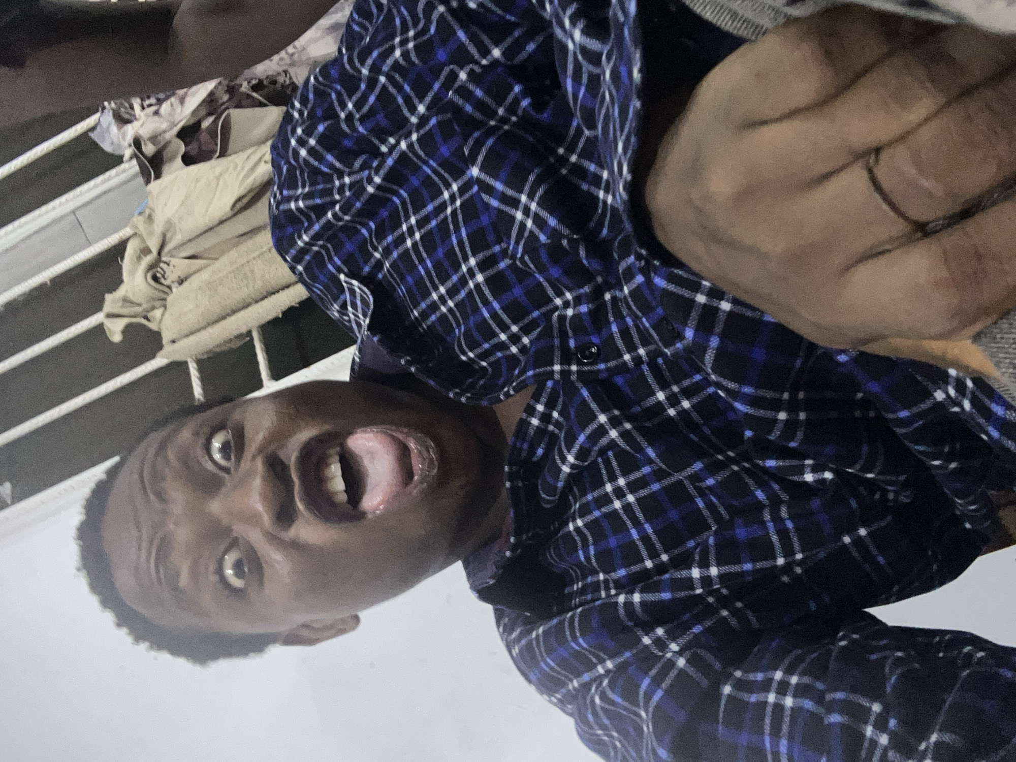 A man with dark skin, wearing a blue and black plaid shirt, is lying on his back with his mouth open. He has short hair and his eyes are wide. A hand wearing a ring is visible near his chest. Clothes are piled on a metal rack in the background, against a white wall. The location is unknown.