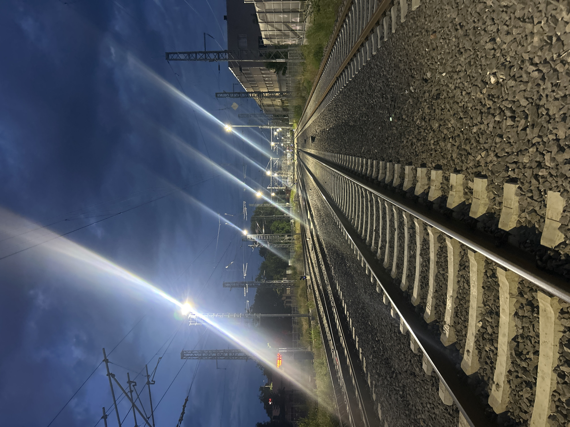 This nocturnal image captures a railway track scene in Plovdiv, Bulgaria, illuminated by numerous artificial light sources under a dark, deep blue sky. The perspective is directed along two parallel sets of railway tracks that recede into the distance, running diagonally from the bottom-left foreground towards the top-right background.
The tracks consist of polished metal rails fastened to light-grey concrete sleepers, which are embedded in a bed of dark grey gravel ballast. Lining the tracks and extending overhead are multiple metallic structures, including poles and gantries, supporting the intricate electrical catenary system for trains.
A series of bright streetlights or floodlights are positioned along the left side of the tracks, casting intense lens flares that streak across the dark sky, adding a dramatic, almost ethereal quality to the scene. These lights provide significant illumination, highlighting the texture of the ballast and the geometry of the tracks.
In the mid-ground on the left, a large, multi-story building with a grid-patterned facade stands partially visible. Beyond the tracks, patches of green vegetation and distant trees are discernible in the dim light. A small, reddish-orange signal light glows softly near the tracks in the mid-ground on the left.
The scene is static, depicting the infrastructure of the railway line without any visible trains or people. The time of day is clearly night or very late twilight, evidenced by the profound darkness of the sky and the prominent role of artificial lighting. The weather appears clear, allowing the sharp visibility of light flares. There is no discernible text visible within the image. The photograph conveys a sense of quiet solitude and the functional beauty of railway engineering at night.