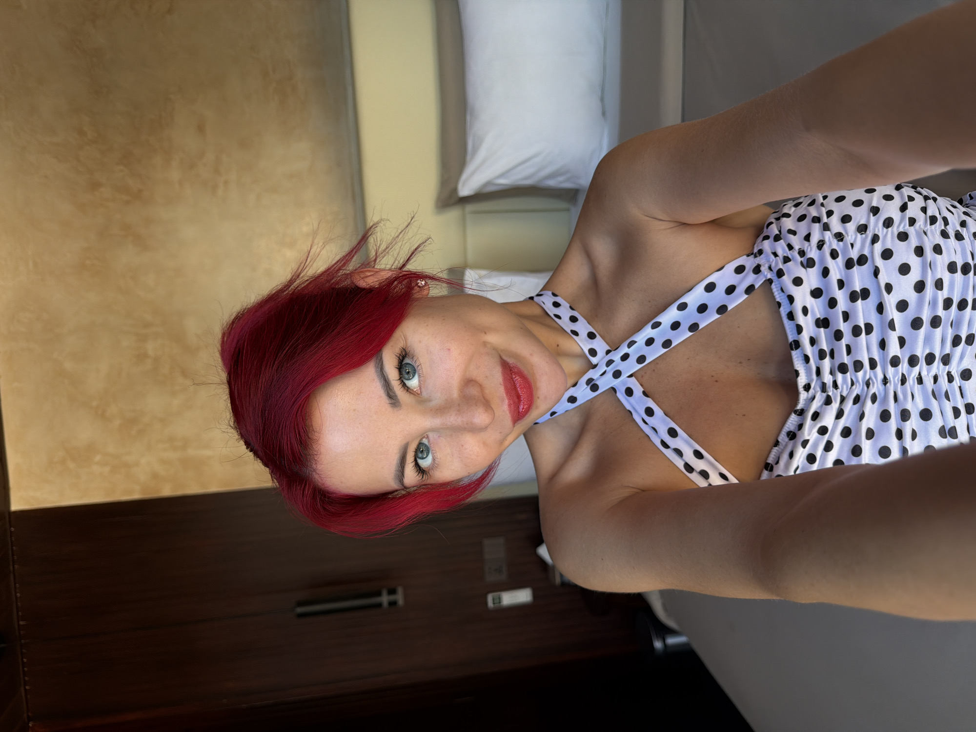 The image shows a medium shot of a woman with bright red hair and blue eyes, looking at the camera. She is wearing a white and black polka dot halterneck top. Her skin has freckles, and she is wearing red lipstick. The background appears to be a hotel room. The wall behind her is textured and light brown, and to her left, there is a dark wood paneling. A bed with white pillows is visible behind her to the right. The woman is positioned in the foreground, holding the camera as if taking a selfie. Based on the context provided by the prompt, this scene is located in Kuta, Indonesia.