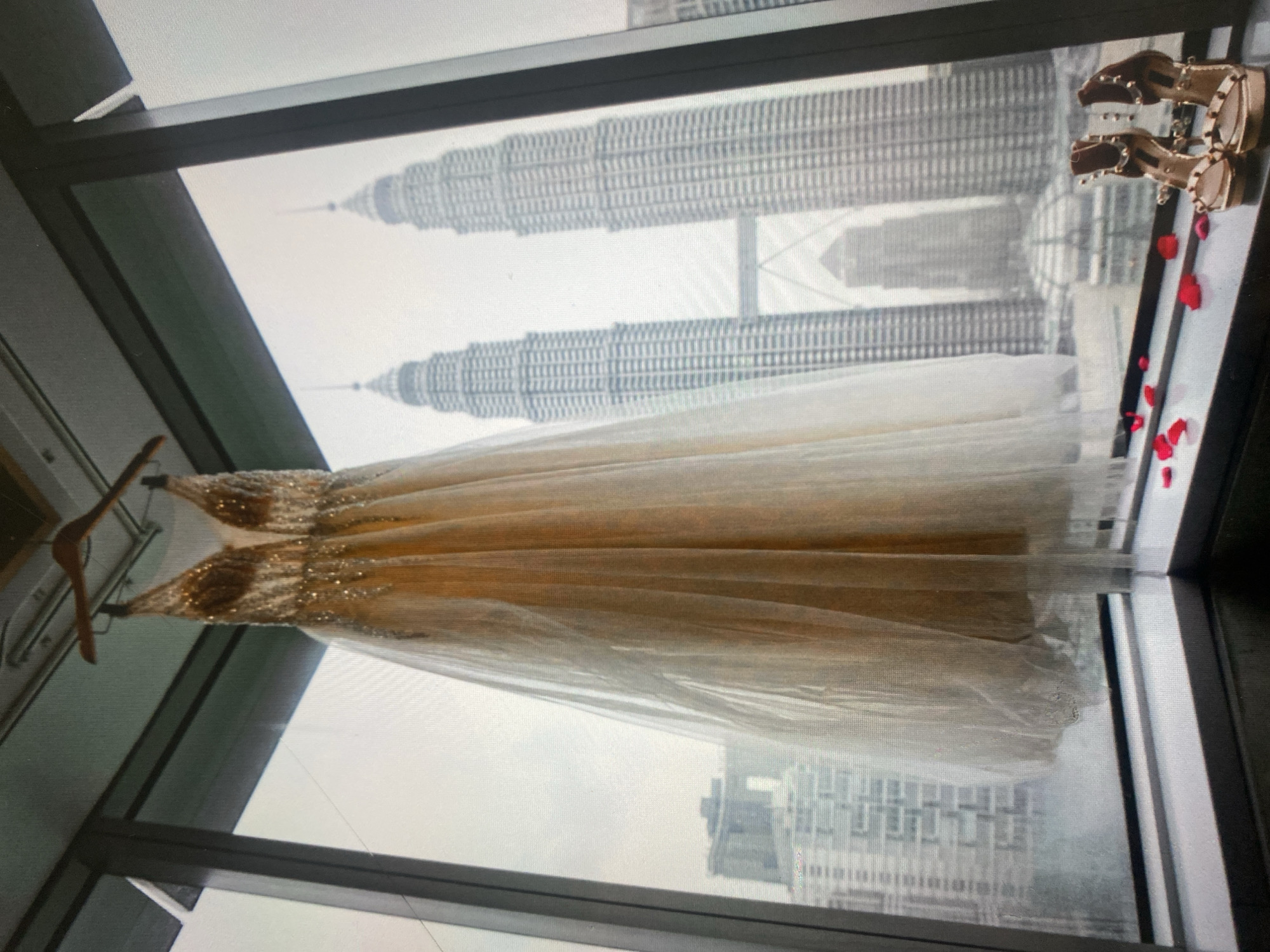 A cream-colored sequined and tulle wedding dress hangs from a wooden hanger in front of a large window, overlooking the city of Kuala Lumpur, Malaysia. The Petronas Twin Towers, a prominent landmark, are visible in the background, shrouded in an overcast sky. Two gold-colored heeled sandals with stud embellishments and rose petals are placed on the windowsill in the foreground. The scene appears to be set in a room with a minimalist aesthetic, emphasizing the view and the bridal attire.
