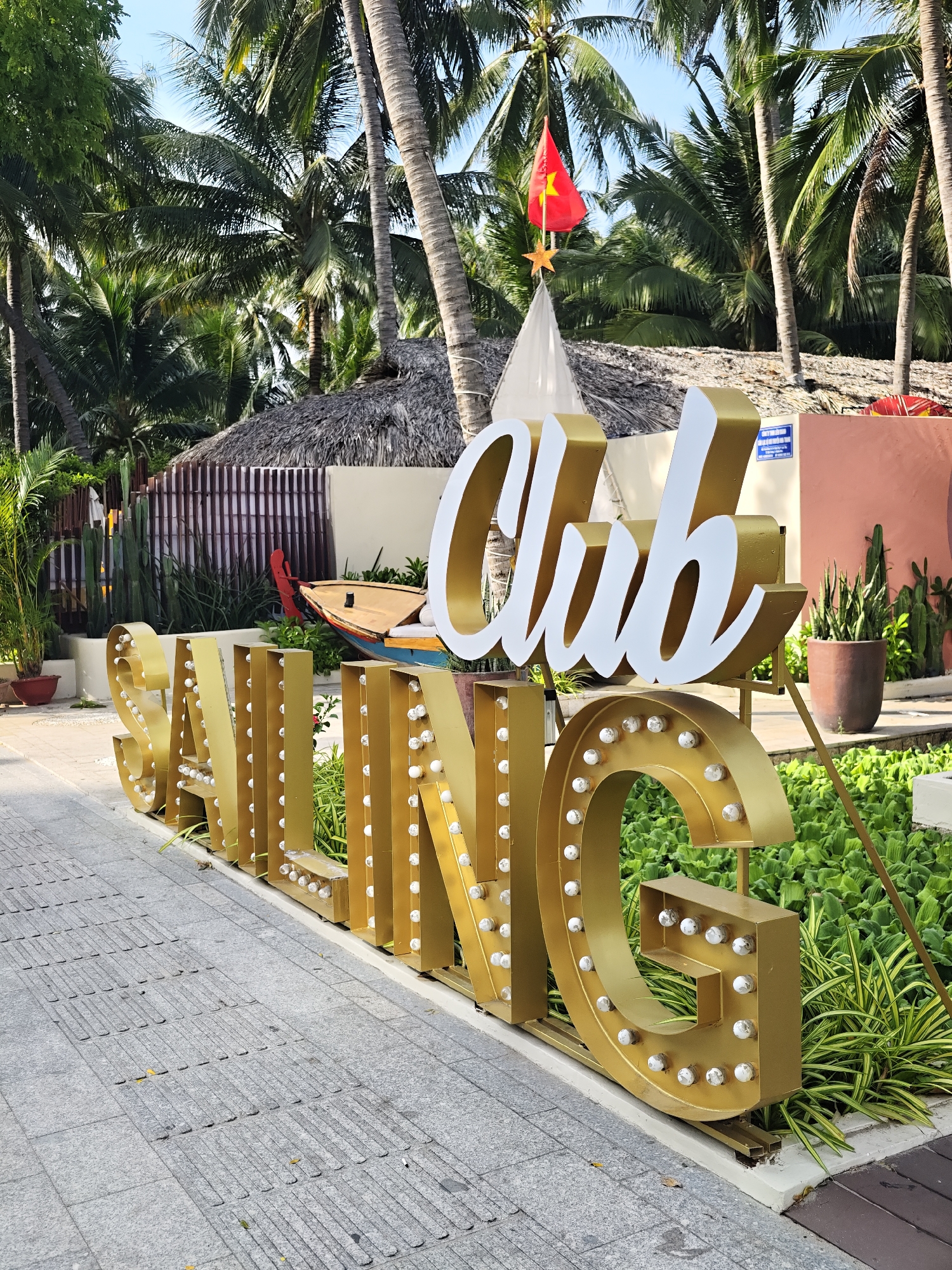The image depicts a brightly lit outdoor scene in Nha Trang Ward, Vietnam, featuring a large, golden marquee sign spelling out "SAILING CLUB." The sign, illuminated with numerous small, white bulbs, is positioned on a paved walkway adjacent to lush green foliage and a body of water. In the background, palm trees and a traditional thatched-roof structure are visible, suggesting a tropical or coastal resort setting. A Vietnamese flag flutters from a flagpole, adding a patriotic element to the scene. The overall ambiance is one of a vibrant and inviting tourist destination.