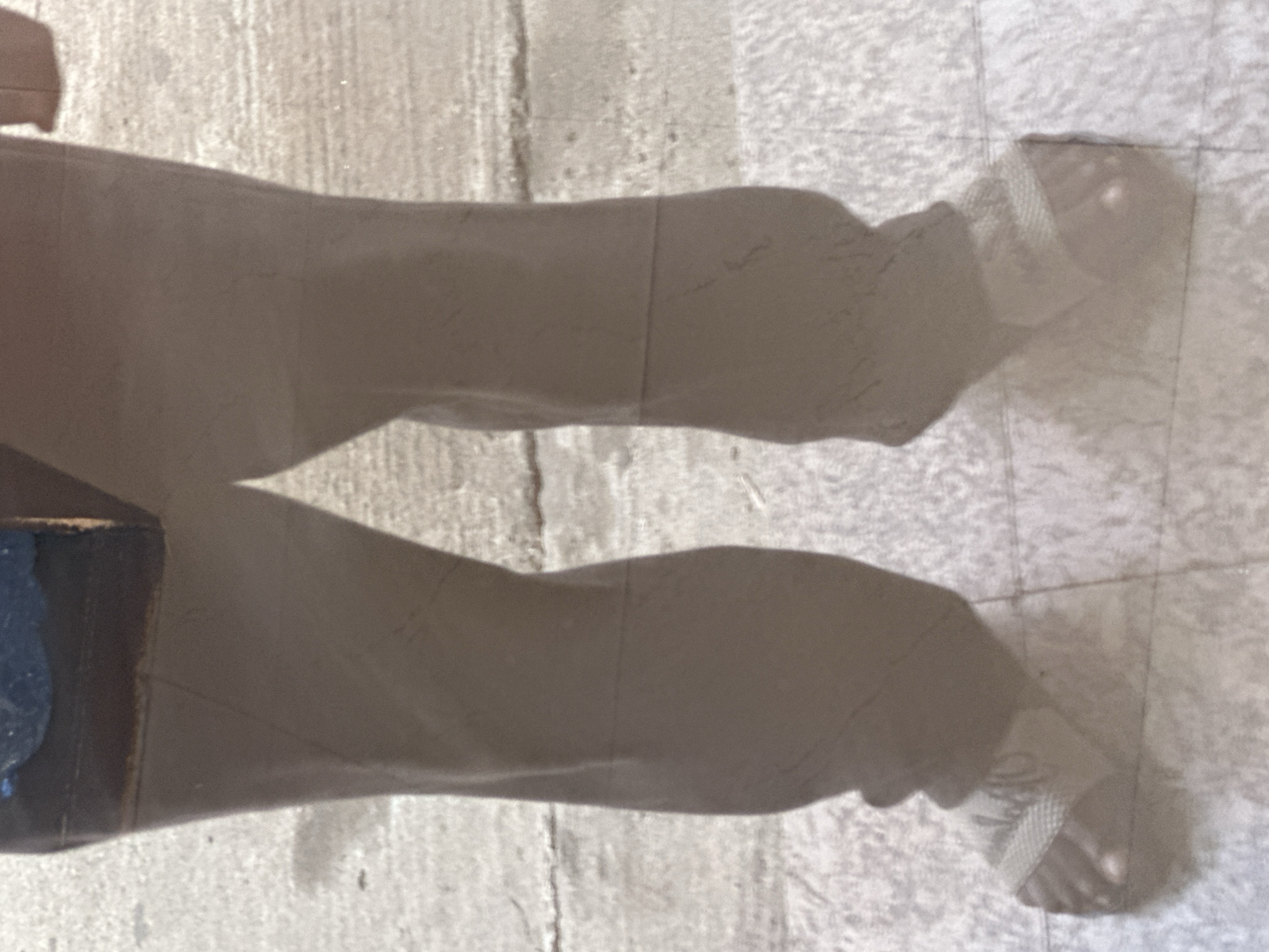 The image shows the lower body of a person wearing dark-colored pants and sandals, standing on a tiled surface. The person's feet are partially visible, with toes peeking out from the sandals. The background appears to be an outdoor setting with a rough, textured surface, possibly a sidewalk or pavement. There are some indistinct shadows and light reflections on the ground. The scene does not provide any specific visual cues to determine the exact location within Gura Top, Nigeria.