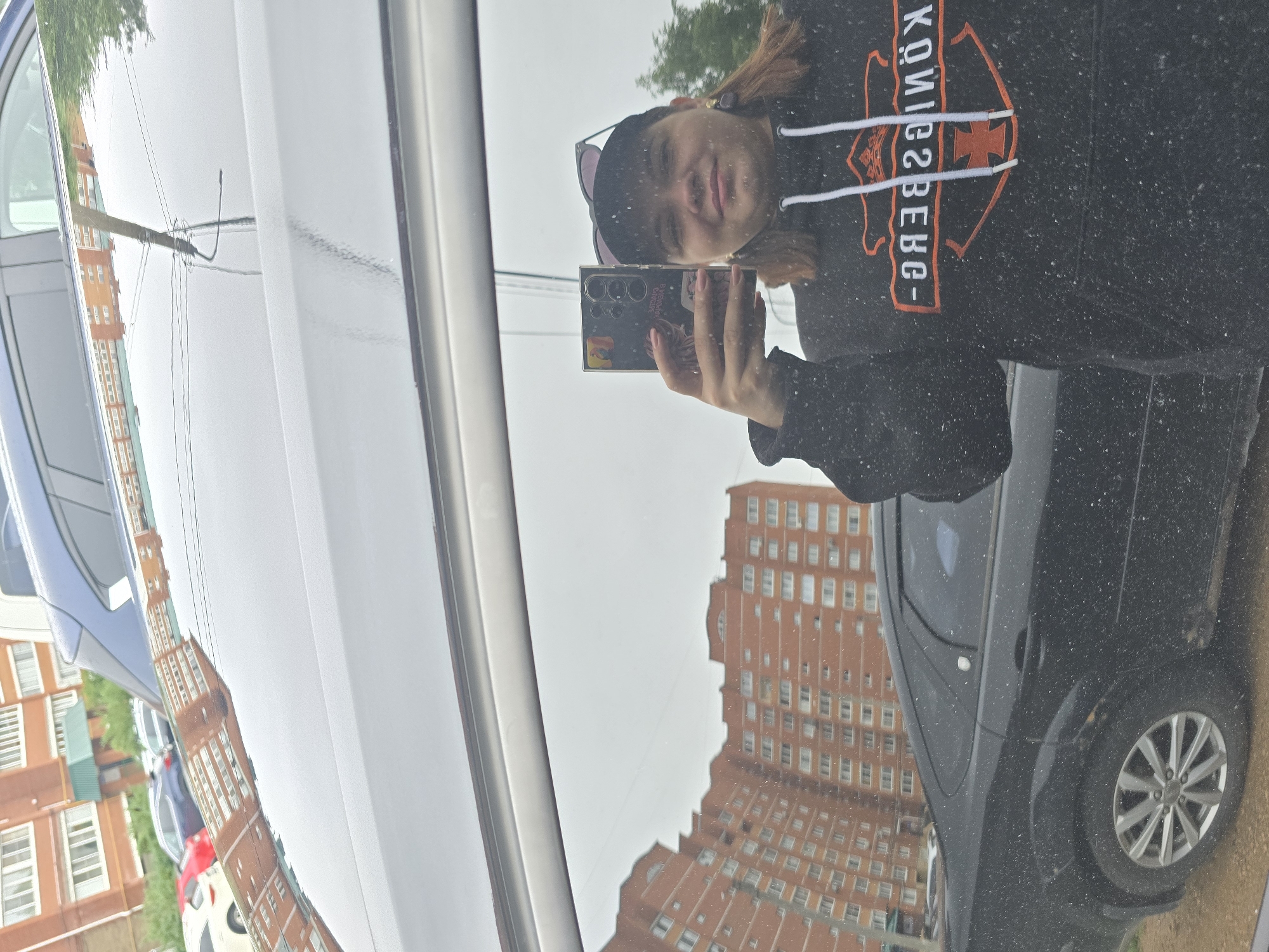 In Chaikovsky, Russia, a person is captured in a reflection on the side of a black car. They are holding a smartphone and taking a selfie. The person is wearing a black hoodie with an orange logo and a baseball cap. In the background, apartment buildings are visible, indicating an urban setting. The sky appears overcast. The car is parked on a dirt or gravel surface.