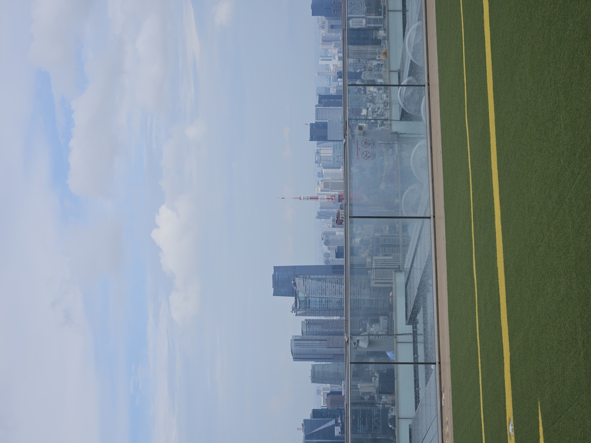 Here is a forensic description of the image:
The image depicts a high-angle view of a cityscape from a rooftop, with the view obscured by a clear glass barrier. The scene is divided roughly into three sections: the sky, the cityscape, and a synthetic green surface. The sky is overcast with patchy, light clouds. The cityscape features densely packed high-rise buildings, with the Tokyo Tower faintly visible. The rooftop is covered with a green synthetic material marked with yellow lines.