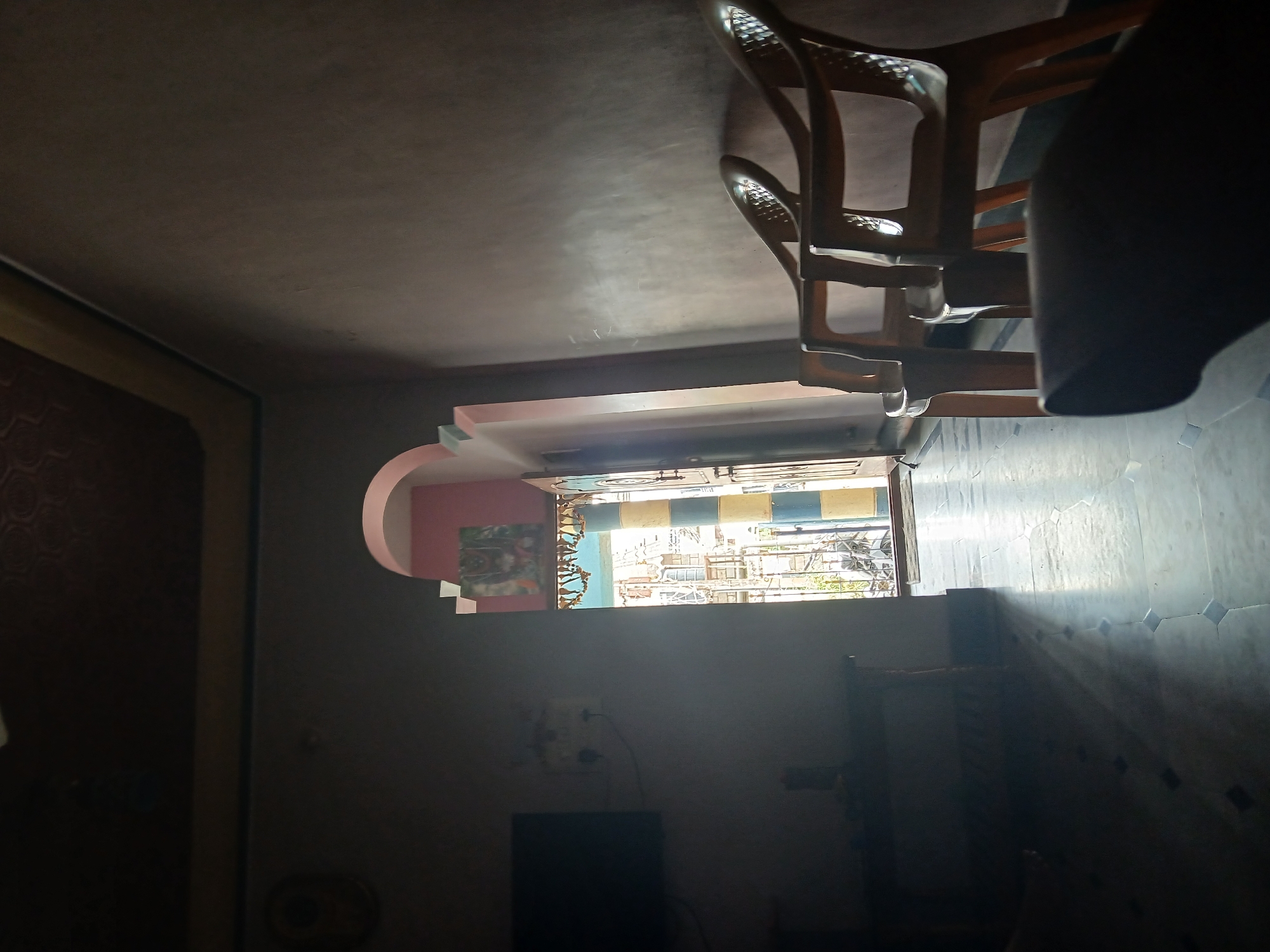 The image shows the interior of a room in Shivamogga, India. A window provides a view of the outside, including structures. In the room, there's furniture like a chair. The walls have a mix of colors and patterns. There are also electronic devices like a TV.