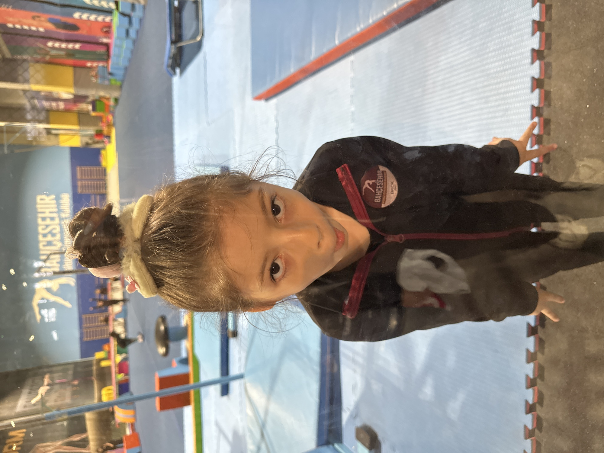 A young girl with brown hair pulled back in a bun stands in front of a glass partition. She wears a dark-colored jacket with red accents, and a patch on the upper right chest reads "BAHCESEHIR BKCK." Her face is turned towards the camera. Reflected in the glass is an indoor sports facility. Gymnastic equipment and colorful padded structures are visible. The wall behind the equipment is blue with white lettering and a logo depicting a gymnast. The floor is covered in blue mats with red markings. Based on the jacket patch, the location is likely in Bahcesehir, Esenyurt, Turkey.