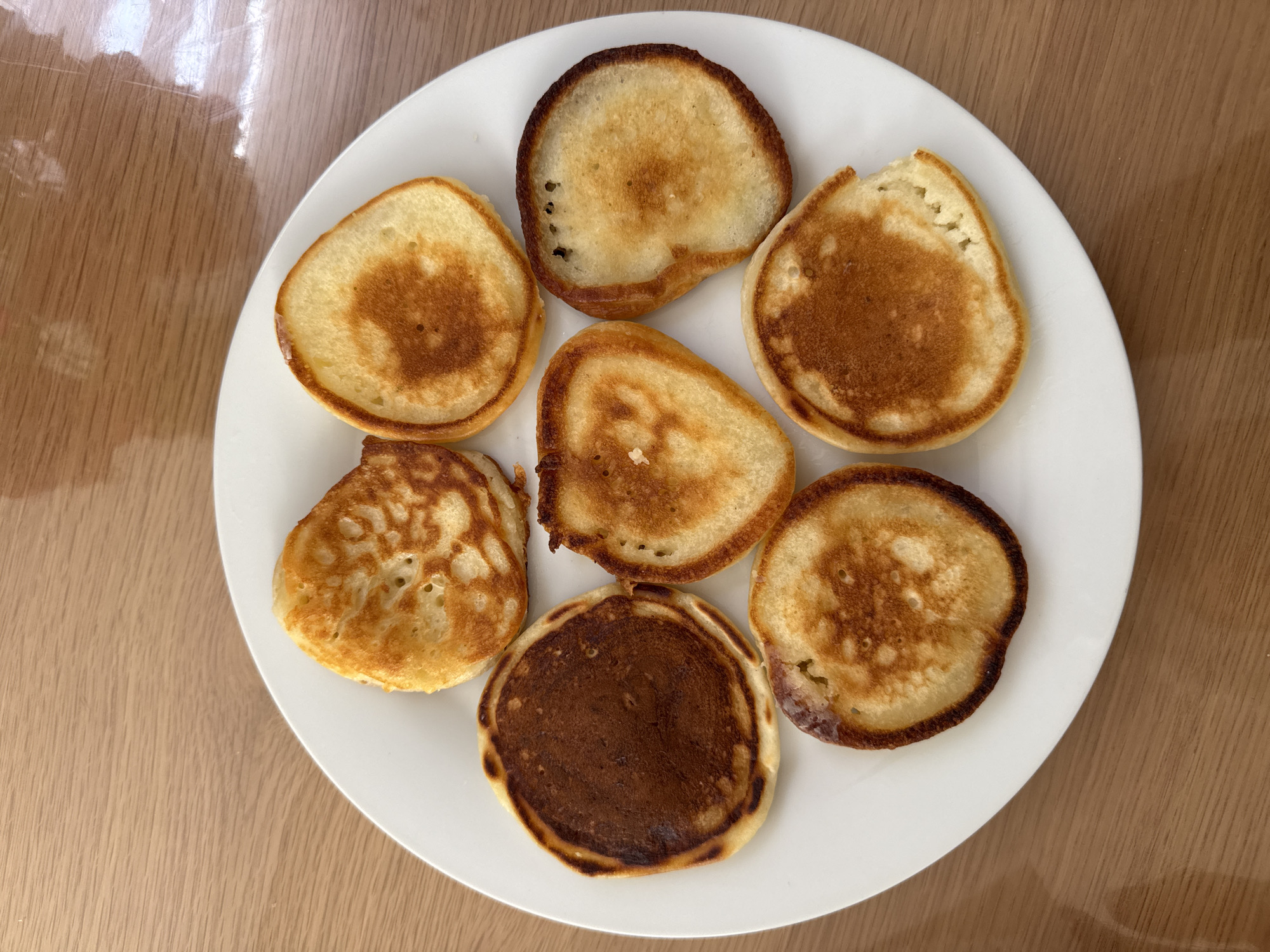 A plate of small, golden-brown pancakes is arranged in a circular pattern on a white plate. The plate is situated on a light brown wooden table. The pancakes vary slightly in color and texture, with some appearing more browned than others. The lighting suggests an indoor setting with natural light coming from the right. There are no people or any other objects visible in the image.