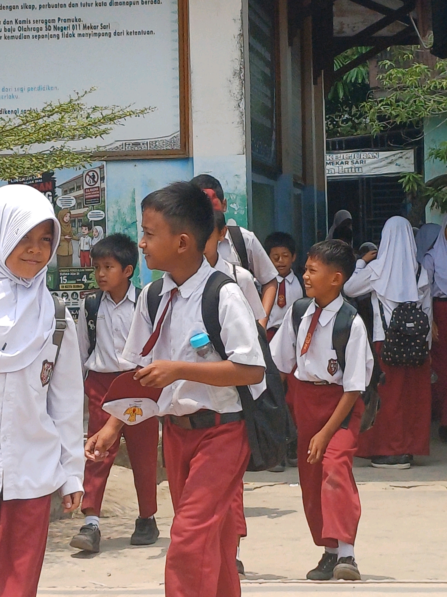 In Dumai City, Indonesia, a group of schoolchildren in uniform are gathered outside a building. The children are wearing white shirts, red pants, and some have black backpacks. Some of the children are looking at each other, while others are looking away. The building appears to be a school, with a mural on the wall. The weather is sunny, and there are trees and plants in the background. The overall scene depicts a typical school day in Indonesia.