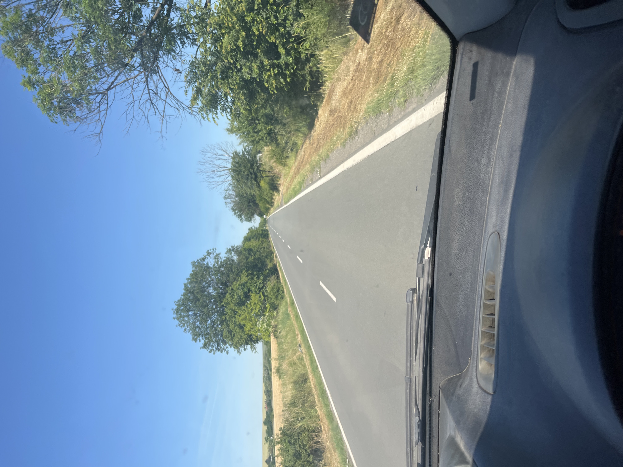 The image depicts a view from the interior of a vehicle looking out onto a rural road on a sunny day. The road is asphalt and has white lane markings. On either side of the road are fields of dry, golden grass or crops, and scattered trees with green foliage. In the distance, the landscape is relatively flat with more trees and possibly some buildings. The sky is clear and blue. The foreground shows the dashboard of the vehicle, including a windshield wiper and part of the steering wheel. There are no people visible in the image. The scene appears to be located in a rural area, likely in or near the city of Ravnets, Bulgaria, given the context provided.