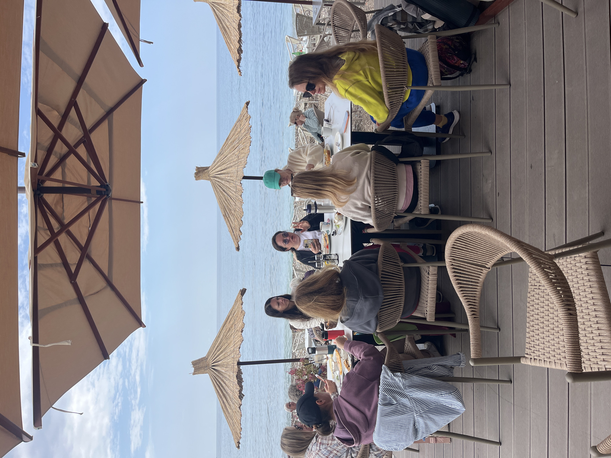 The image shows a group of people dining outdoors at a restaurant in an unknown city. They are seated at tables with chairs, positioned on a wooden deck overlooking the sea. Each table has an umbrella providing shade from the sun. The diners are dressed in casual clothing and are engaged in conversation and eating. The sky is visible in the background and is blue.