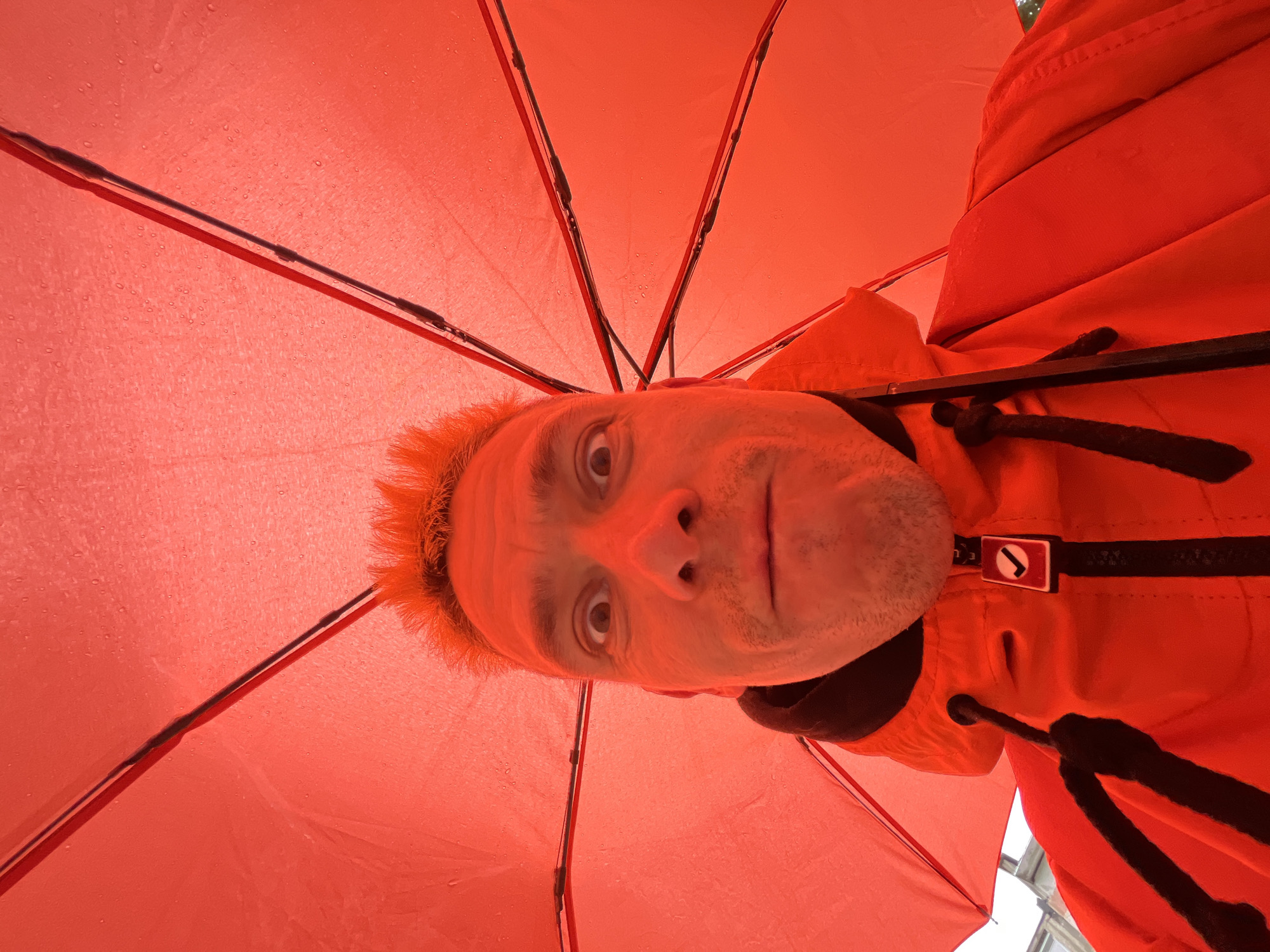A man is holding an open red umbrella over himself. He is looking directly at the camera with a surprised expression. He has short, spiky red hair and a mustache. He is wearing a red jacket with black drawstrings and a black hoodie underneath. The umbrella is covered in raindrops, indicating it is raining. The background is blurry and suggests an outdoor urban environment, though no specific landmarks of Chaikovsky, Russia are visible.