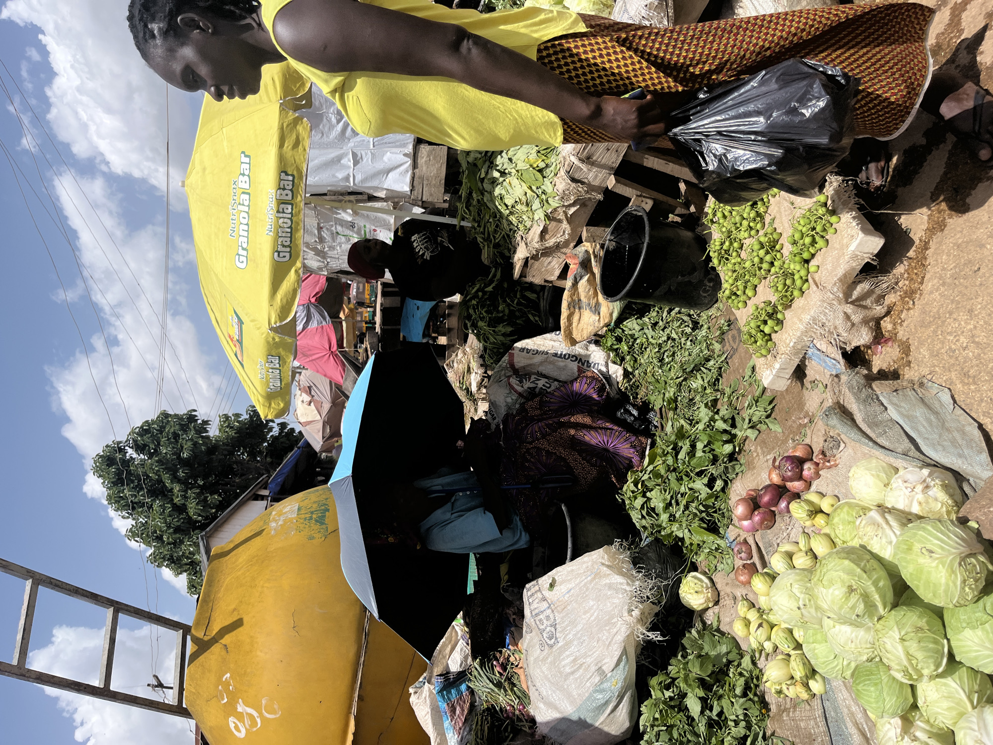 This media file depicts an outdoor market scene in Jos, Nigeria. The primary subjects are individuals and a variety of fresh produce. In the foreground, a person wearing a bright yellow top and a patterned skirt is holding a black plastic bag. Next to them, on a rough, burlap-like surface, are piles of green vegetables and a collection of small, round green fruits or vegetables. Further to the right, there are several heads of cabbage and a cluster of red onions.
In the mid-ground, an individual wearing a red cap and a dark t-shirt is visible, as well as other market-goers and stalls. A large yellow umbrella with the text "NutriSnax Granola Bar" printed on it is prominent, providing shade. Another umbrella, this one a vibrant blue and black, is also visible, partially covering a person dressed in blue. Various sacks and wooden crates are used as displays for the produce.
The setting appears to be a bustling marketplace with elements suggesting a sunny day, indicated by the clear blue sky with scattered clouds and the shadows cast. The overall atmosphere suggests daily commerce and the sale of fresh, locally sourced food items. The composition highlights the vibrant colors of the produce and the people's clothing against the earthy tones of the market.
