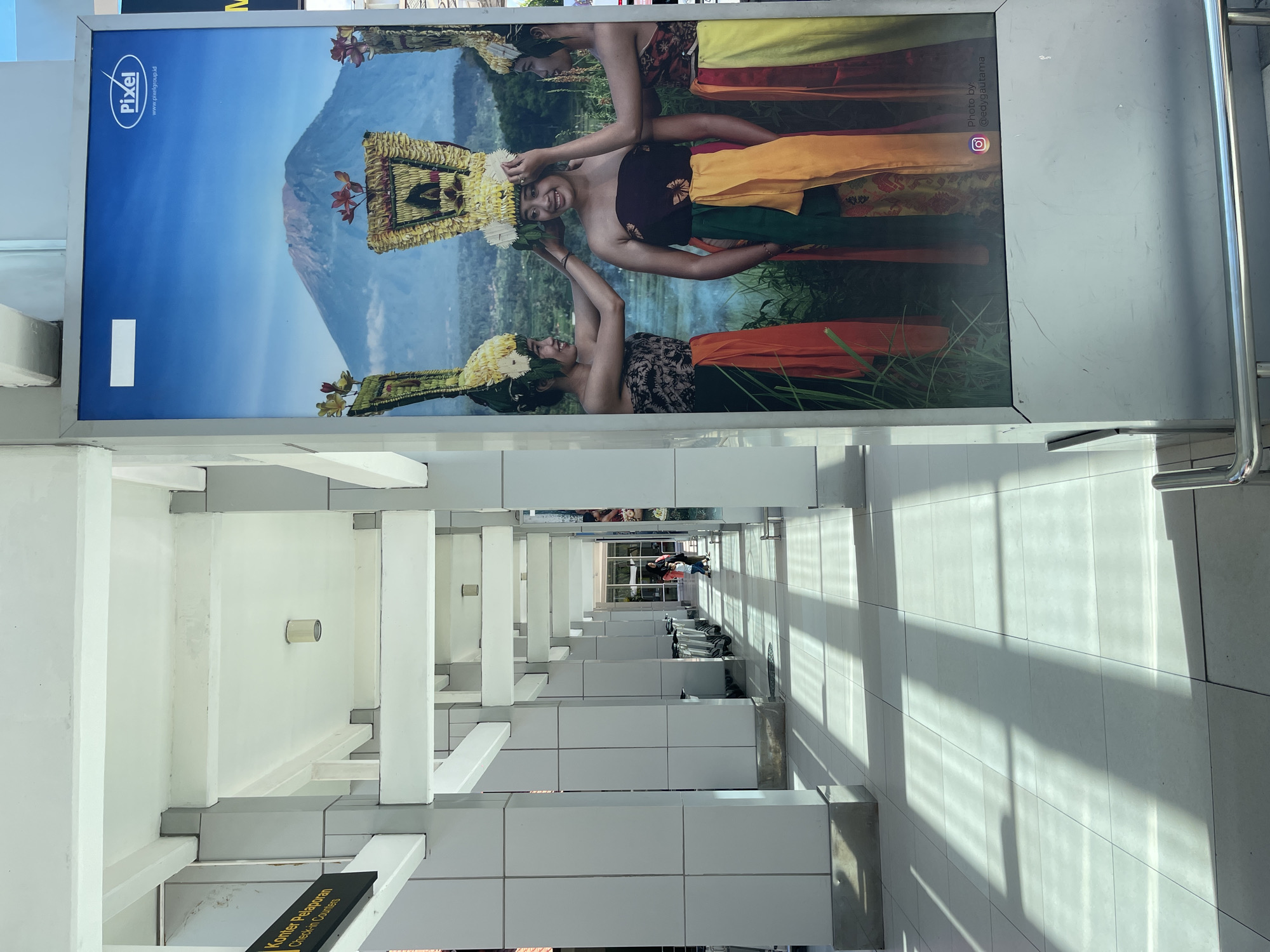 The image depicts an outdoor covered walkway at what appears to be an airport or transportation hub in Kuta, Indonesia. The walkway is lined with white pillars and a white ceiling, with natural light filtering in and casting distinct shadows on the tiled floor. In the foreground, a large digital advertisement is prominently displayed on the right. This advertisement features a vibrant image of three young women in traditional Balinese attire, adorned with elaborate headdresses made of flowers and gold. They are positioned in front of a lush green landscape and a majestic mountain in the background. The advertisement also includes the "Pixel" logo and a website address. On the left side of the walkway, a dark sign with white text reads "Konter Pelaporan" (Reporting Counter) and "Check-in Counters." Several rows of luggage carts are visible further down the walkway, and a few people are walking in the distance. The overall impression is one of a clean, modern public space with a cultural advertising element.