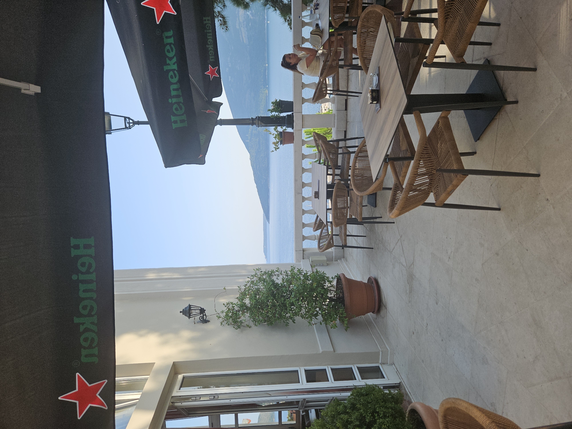 The image shows an outdoor cafe with a view of the sea in Herceg Novi, Montenegro. Several tables and chairs are set up on a paved terrace. In the foreground, a table with woven chairs is visible. In the middle ground, there are more tables and chairs, with a railing and the sea in the background. A woman is seated at one of the tables, holding a glass. The cafe has umbrellas with the Heineken logo. The architecture suggests a building with white facade and columns. The overall scene depicts a leisurely setting by the water.