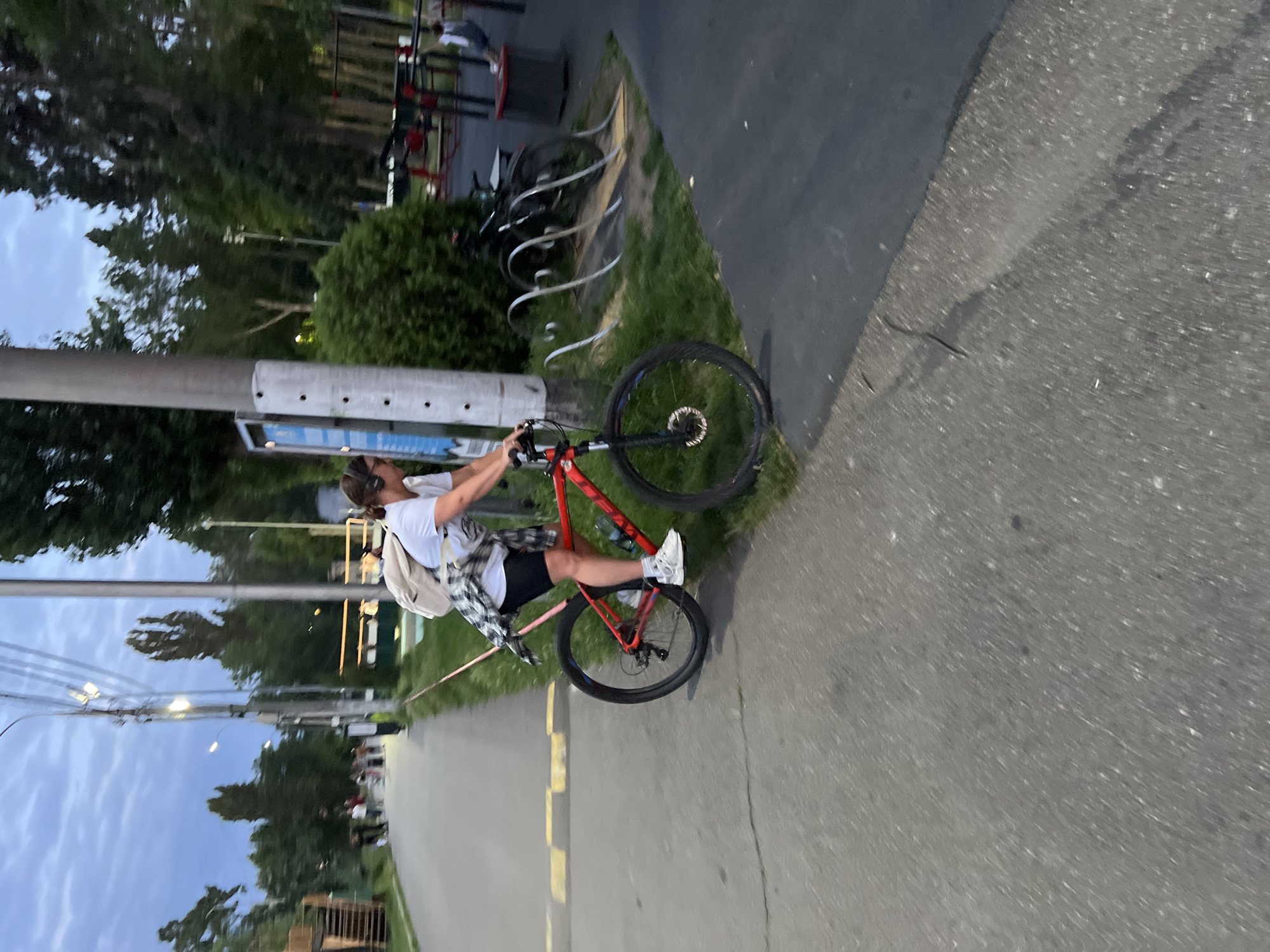 A woman is riding a red bicycle on a paved path in a park in Krasnodar, Russia. She is wearing a white t-shirt, black shorts, white sneakers, and headphones. A light-colored backpack is slung over her shoulders, and a checkered shirt is tied around her waist. The bicycle has a water bottle cage attached to the frame. In the background, there are trees, a street light, a bicycle rack with several parked bicycles, and an outdoor fitness area with red and black equipment. The sky is overcast, indicating it is likely early morning or late evening. The path is bordered by grass. Other people are visible in the distance, walking on the path.