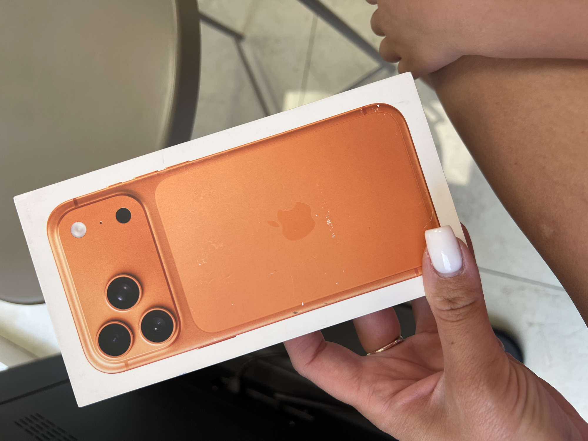 A close-up image shows a person holding an iPhone box in a bright, daytime setting. The central subject is a female hand with well-manicured, white, square-shaped nails, wearing a gold ring on the index finger, holding a white smartphone box.
The box prominently displays a large, glossy image of the back of an orange or gold-colored iPhone. This depicted phone features a triple-camera system, with three dark circular lenses arranged in a triangular cluster, accompanied by a small flash unit. The iconic Apple logo is visible in the center of the phone's depicted back. Some minor smudges or marks are present on the illustrated phone surface on the box.
Portions of the person's tanned arm and leg are visible in the background, with a noticeable patch of lighter skin on the upper arm/leg. The background itself is softly blurred, showing indistinct shapes and muted colors of brown and grey, suggesting an indoor or covered outdoor environment like a cafe, restaurant, or balcony. A dark object, possibly furniture, is partially seen in the lower left corner. The scene takes place in Budva, Montenegro. No discernible text is visible on the box, only the Apple logo.