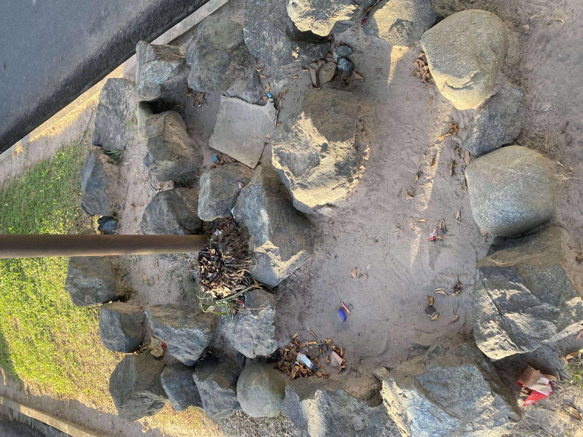 The image depicts a patch of ground surrounded by large rocks, with a metal pole in the center. The ground appears to be sandy and dry, with scattered leaves and a few pieces of litter, including a blue bottle cap and red and white wrapper. In the background, there is a grassy area and a paved road. The scene appears to be in a public outdoor space, possibly a park or roadside area. There are no people visible in the image. The lighting suggests it is daytime, with the sun casting shadows. The overall impression is one of a somewhat neglected outdoor area.