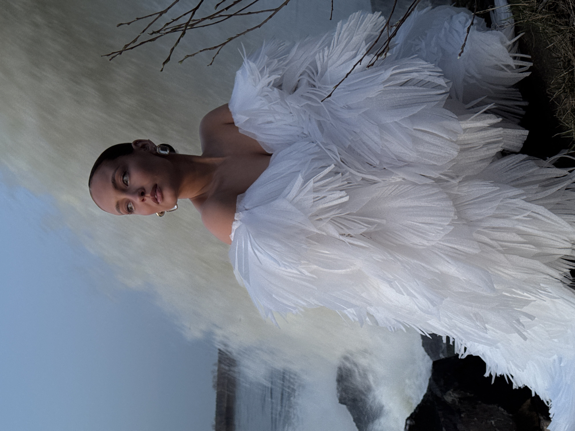 A woman with dark hair pulled back is positioned in front of a waterfall. She is wearing large, square earrings and a white, feathery garment. Bare branches are visible at the top of the frame. The location appears to be outdoors, alongside a waterfall. There is no specific evidence to confirm the image was taken in Chernushka, Russia.