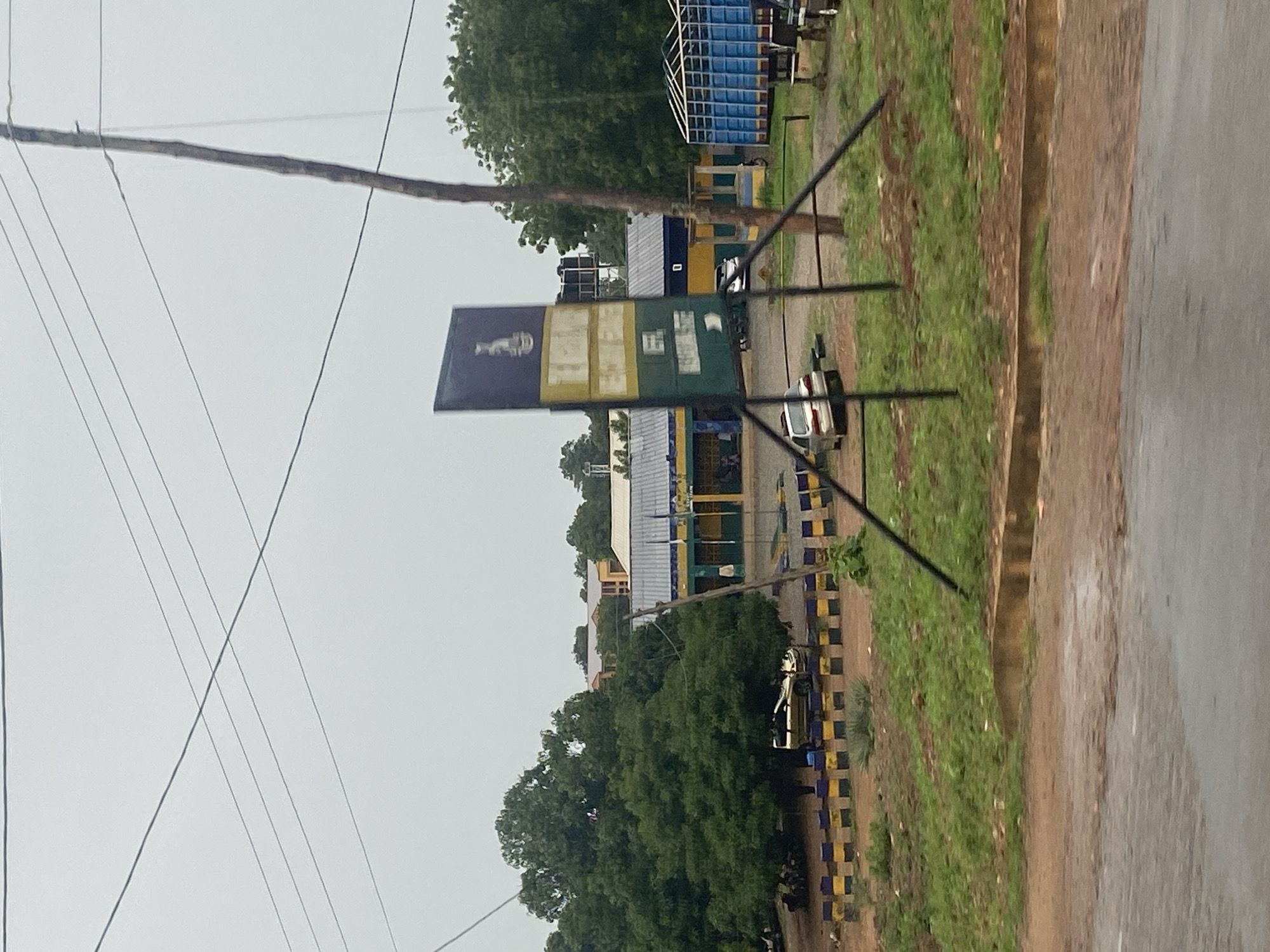 The image depicts a street scene in Keffi, Nigeria, under an overcast sky. In the foreground, a dirt and asphalt road is visible, with a concrete curb separating it from a grassy area. Several yellow and black bollards line the edge of the grassy area.
Further back, a building with a grey metal roof and blue and green painted walls is present. The building appears to be a public or institutional facility, possibly a police station given the presence of a signpost with an emblem. This signpost is a tall, rectangular structure with multiple panels, some of which are yellow and green, and bears a crest at the top.
Several vehicles are parked in front of the building, including a silver sedan, a gold SUV, and a blue truck with a white trailer. A few people are visible near the building's entrance, appearing to be standing or walking.
The background is dominated by lush green trees, indicating a semi-urban or suburban setting. Power lines crisscross the sky above the scene. The overall atmosphere is muted, consistent with the cloudy weather.