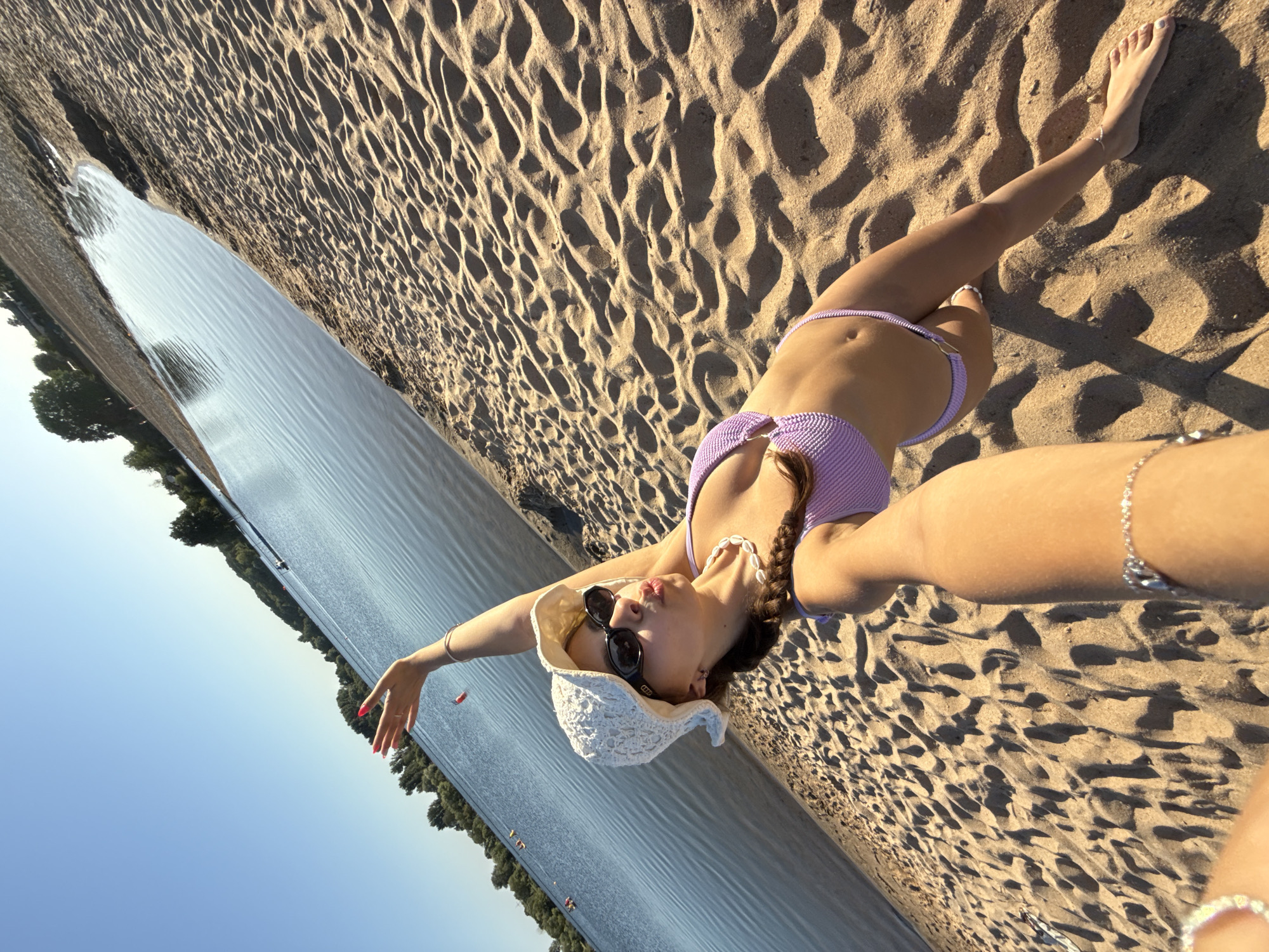 The image shows a young woman on a sandy beach next to a body of water. She is wearing a white crochet hat, sunglasses, and a light purple bikini. She is holding up her right arm and her left arm is holding the camera, taking a selfie. Her left leg is extended, and her right leg is bent. She is wearing a shell necklace and a bracelet on her left wrist. The beach is covered in sand with visible footprints and ripples. The water is calm and reflects the blue sky. Trees line the far edge of the water, and the sky is clear and blue. The location appears to be the Kinzigsee in Langenselbold, Germany, based on the general landscape of a sandy beach adjacent to a lake, typical of recreational areas in that region.