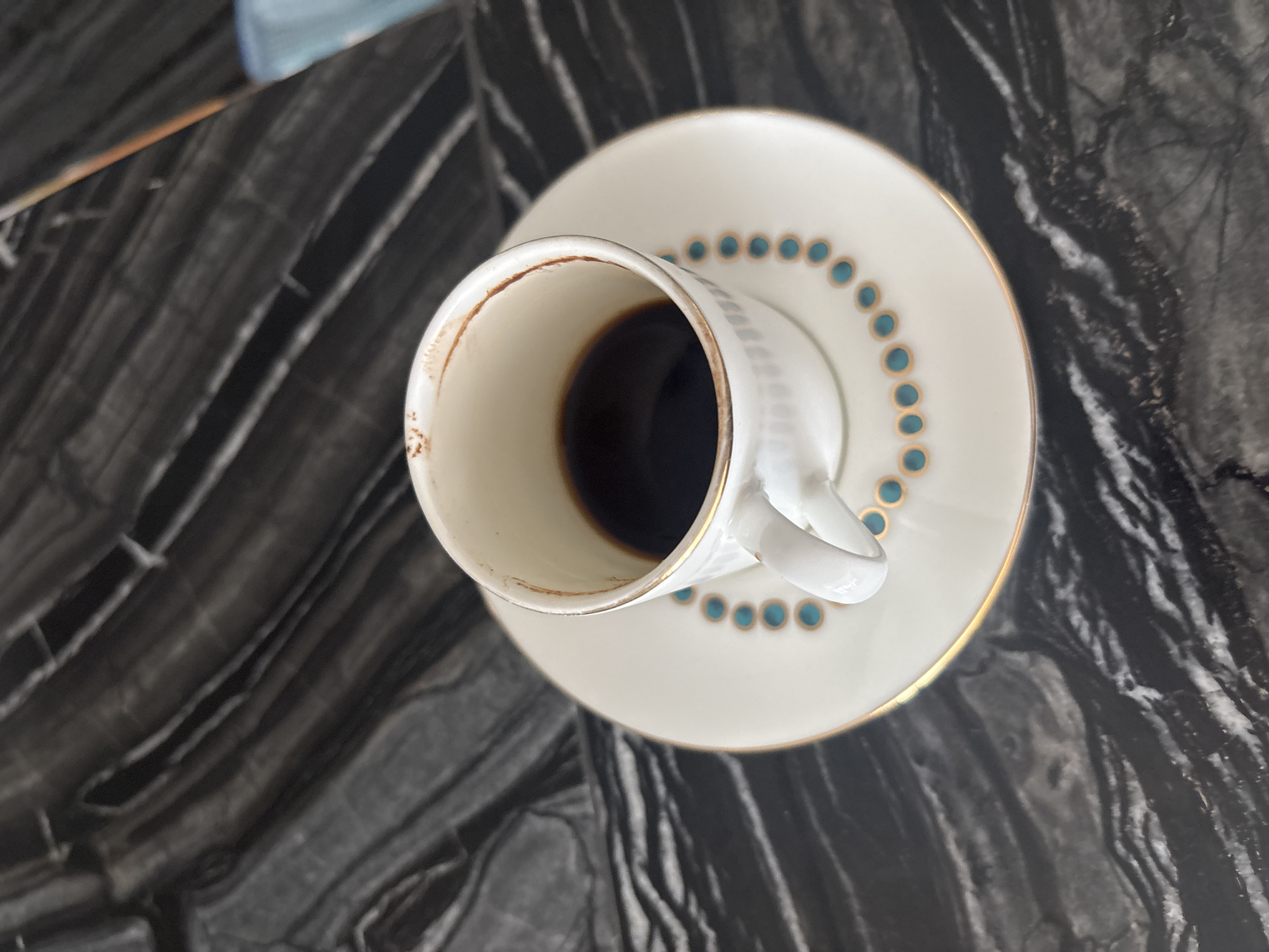A close-up, overhead shot shows a white porcelain cup and saucer with gold trim and a turquoise dot pattern. The cup contains a small amount of dark liquid, likely coffee. The set rests on a black and gray marbled surface, possibly a countertop or table. The background is blurred, suggesting an indoor environment. There is no indication of the specific location within Dubai, United Arab Emirates.