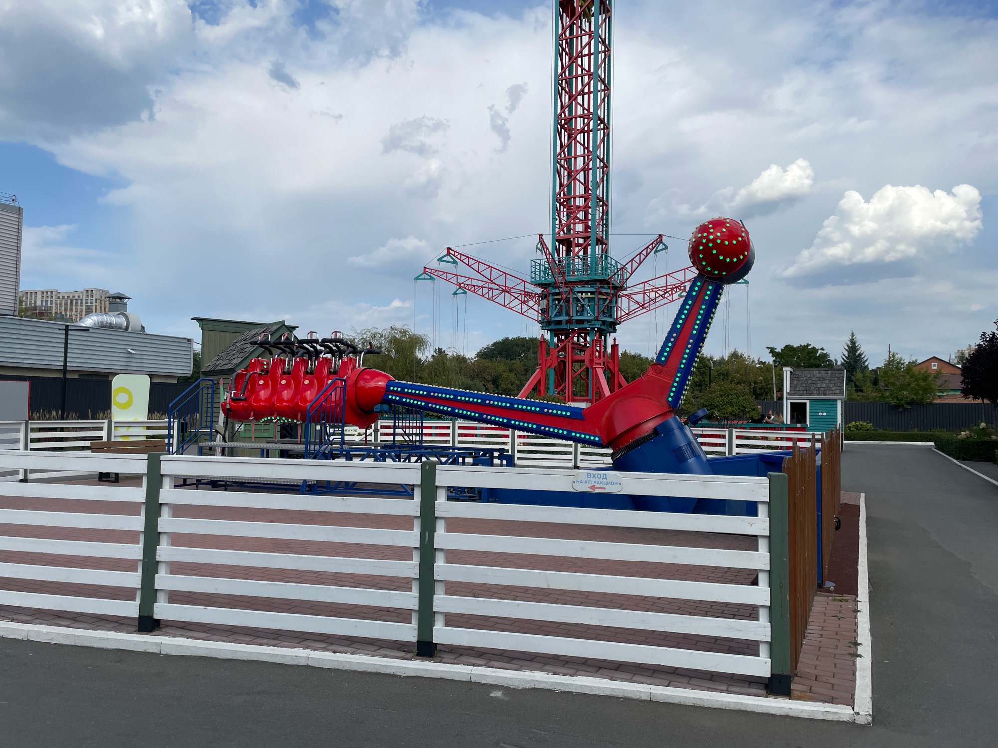 The image shows an amusement park ride, possibly a swing ride or a drop tower, at an amusement park in Omsk, Russia. The ride is red and blue with a tall red and teal tower structure. There are several red seats suspended from the structure. The ride is surrounded by a white wooden fence. The ground is paved with asphalt. The sky is cloudy. In the background, there are some buildings and trees. A sign in Russian indicates "Entrance to the attraction".