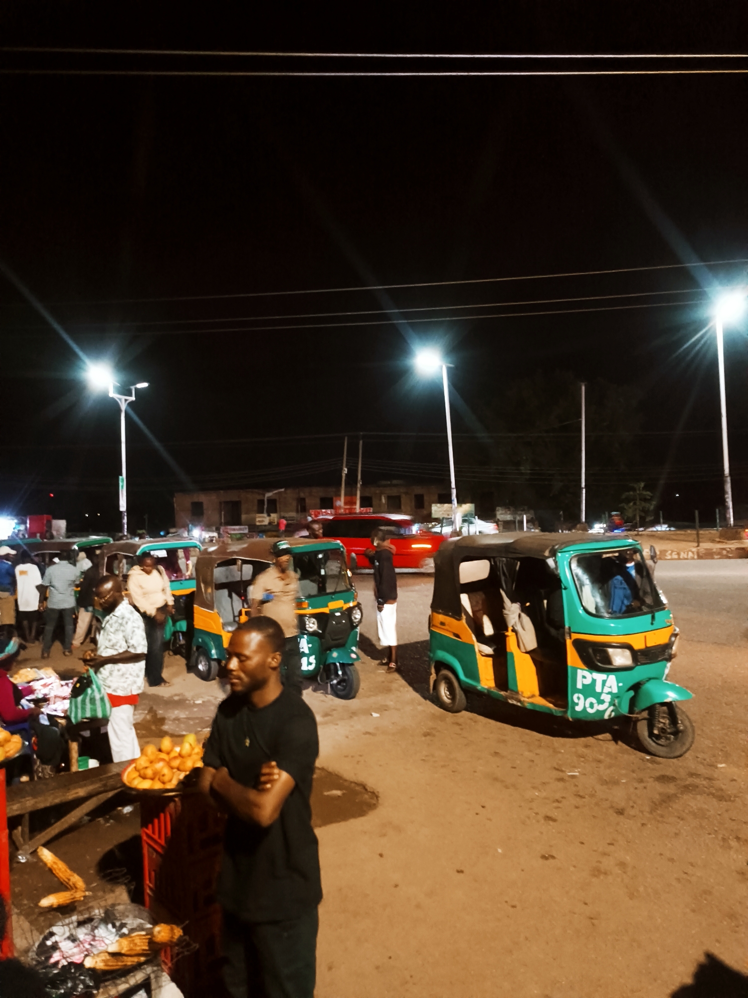 The image captures a night scene in Bukuru, Nigeria, under the bright illumination of streetlights. Several auto-rickshaws, also known as "Keke Napep", are parked along the roadside, painted in a combination of green and yellow. People are gathered around what appears to be a street food vendor, with a man standing in the foreground, arms crossed. A red vehicle passes on the road in the background. The setting is a typical urban area, with buildings visible in the distance.
