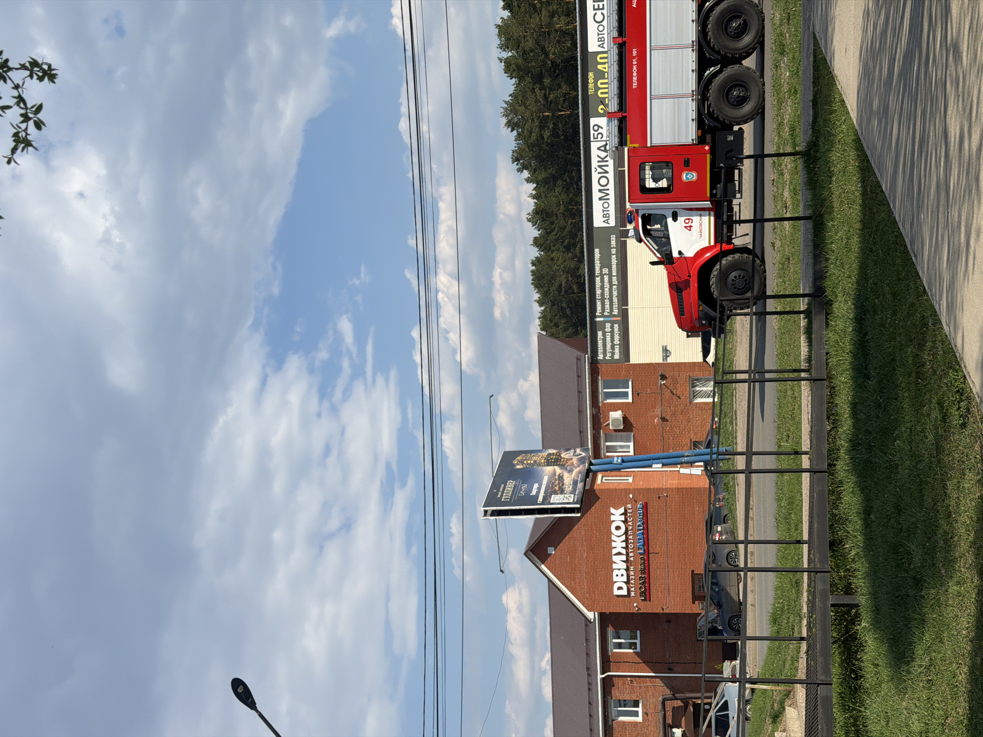 The image shows a street scene in Chaikovsky, Russia. A red and white fire truck with "49 Чайковский" (Chaikovsky) printed on its side is parked along a sidewalk. A building with a sign reading "ДВИЖОК МАГАЗИН АВТОЗАПЧАСТЕЙ" (Dvizhok Auto Parts Store) and "LECAR Store ADA Деталь" (Lekar Store Ada Detail) is also visible. A billboard displaying a building and the word "ГУЛЛИВЕР" (Gulliver) is attached to the building. The scene also includes a light post, utility lines, a fence, trees, and a blue sky with white clouds. A sign on the fire truck reads "АВТОМОЙКА 592-00-40 ABTOCEL" (Car Wash 592-00-40 Auto Cell), listing various auto services.