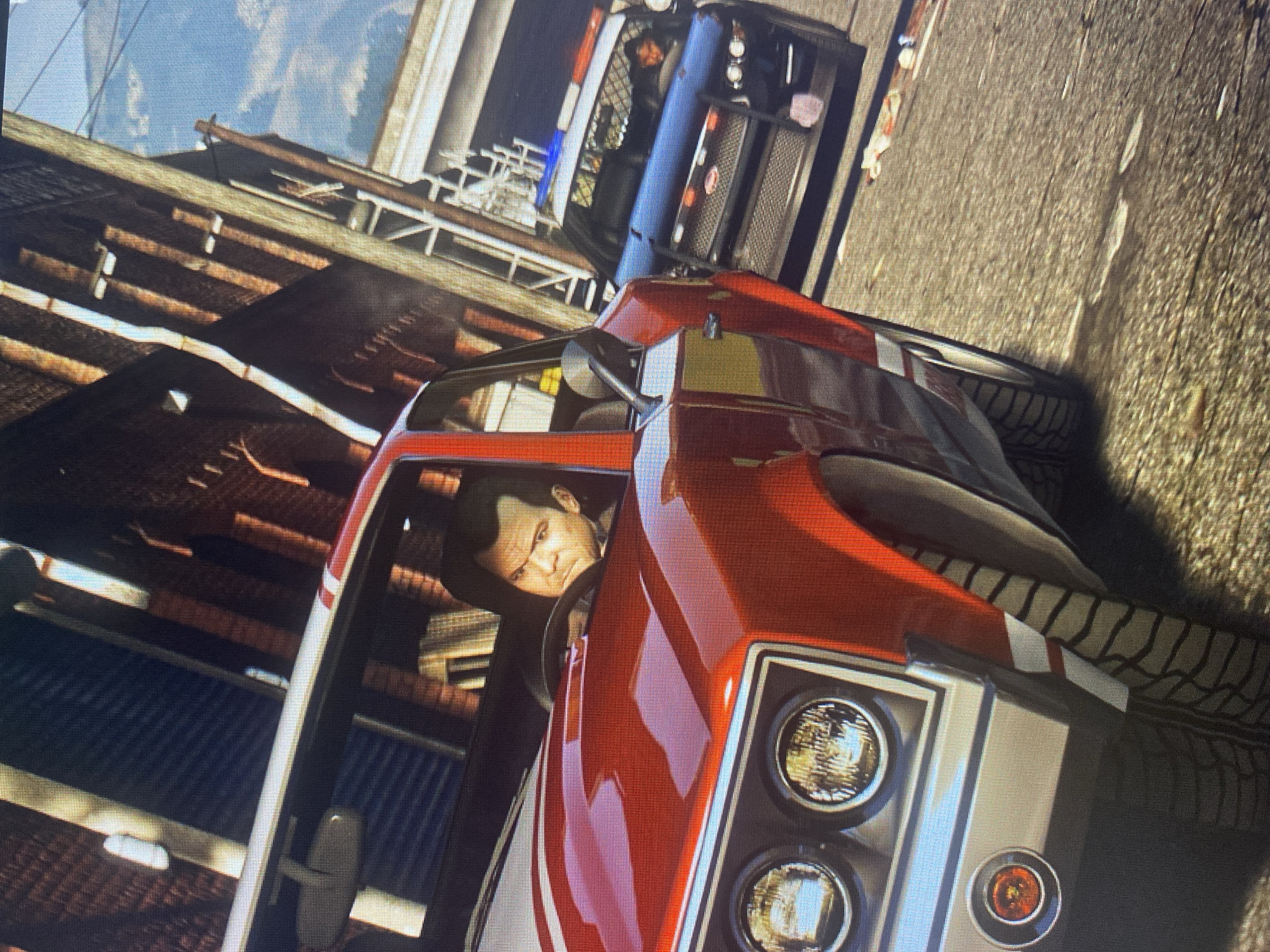 In the city of Unknown, Unknown, a red sports car is driving down a street lined with brick buildings. The driver of the sports car, a male with short brown hair, is looking intently forward. A police car is in pursuit behind the sports car. The police car has its lights on and appears to be gaining on the sports car. The scene is set during the daytime, with clear skies.
