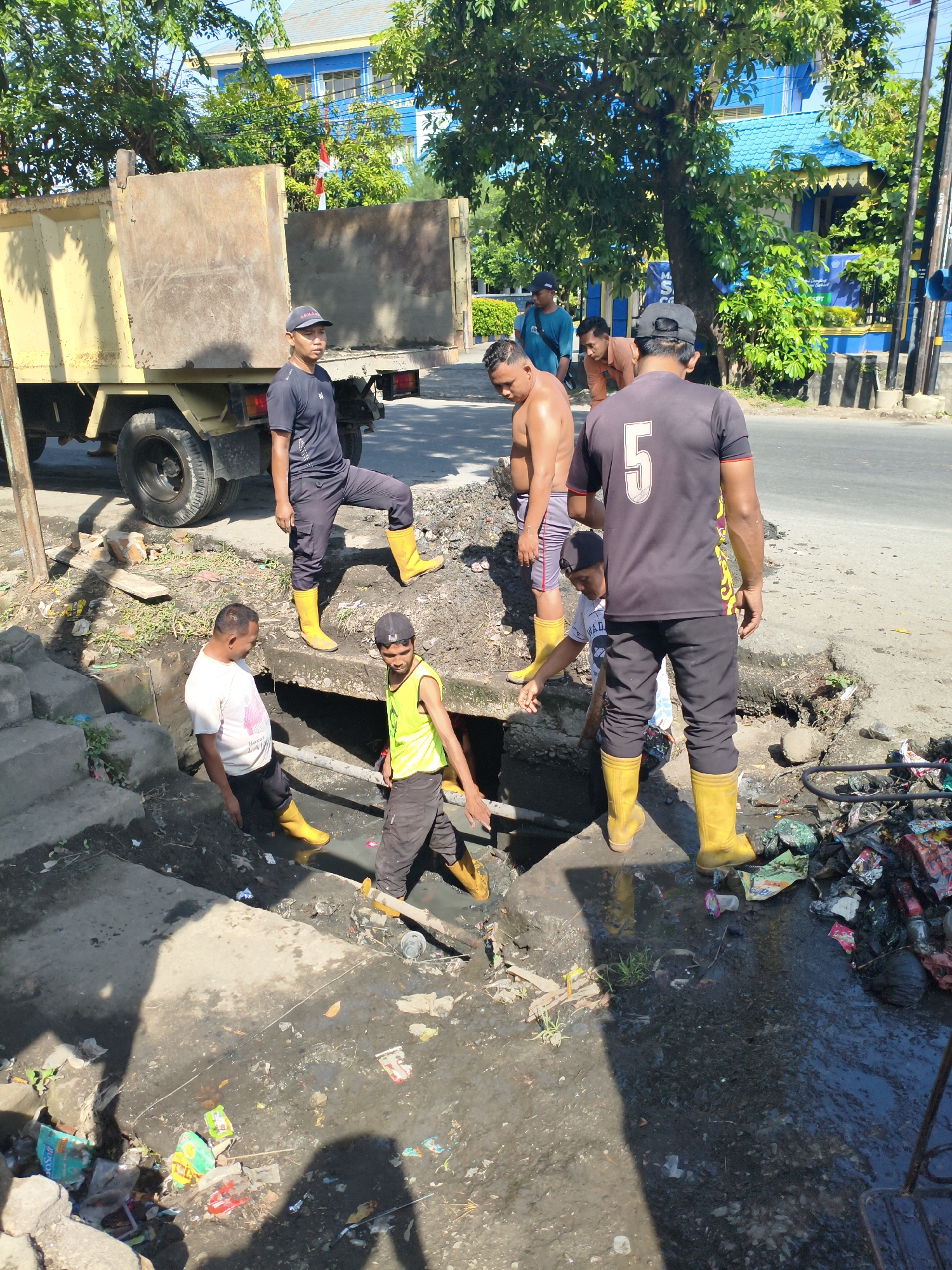 A group of five men are cleaning a clogged drainage ditch in a city. The ditch is filled with murky water and debris. The men are wearing work clothes and yellow rubber boots. One man is in the ditch, reaching into the pipe, while another stands on the edge, looking down. The other three men are standing on the side, observing or preparing to assist. A yellow dump truck is parked nearby, suggesting it may be used to haul away the collected debris. The background shows a street, trees, and buildings, including a blue building with a red and white flag, consistent with a location in the city of Tebing Tinggi, Indonesia. The scene depicts an urban sanitation effort in a tropical climate, indicated by the lush greenery and bright sunlight.