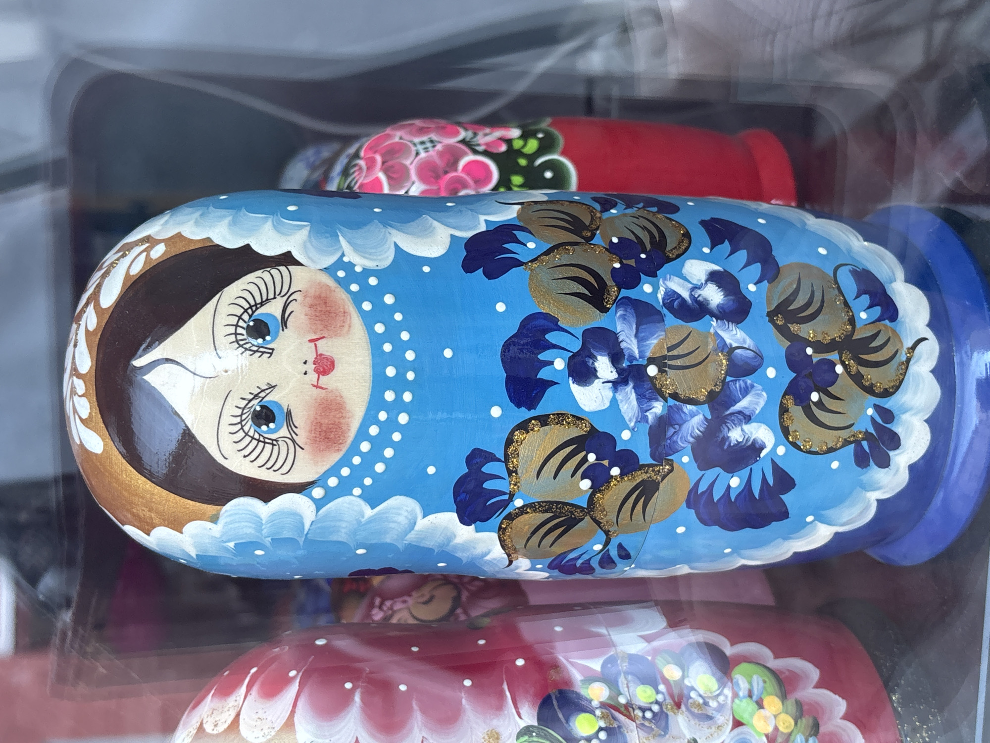 This image, captured in Tallinn, Estonia, displays a prominently featured, brightly painted Matryoshka doll. The doll is centrally positioned, presenting a light-skinned face with blue eyes, long black eyelashes, rosy cheeks, and red lips. Dark brown hair is visible beneath a golden headband adorned with white floral motifs. The doll's head is framed by a light blue headscarf-like design with white scalloped edges. Its upper body is painted a vibrant sky blue with small white polka dots. The lower section of the doll's dress features intricate floral and leaf patterns in dark blue, lighter blue with white accents, and metallic gold with black details, frequently highlighted with gold glitter. The base of the doll is a darker shade of blue, also edged with white scalloping.
To the left and right, partially visible Matryoshka dolls flank the central figure. The doll on the left is predominantly red with white and pink floral designs. The doll on the right is also red, featuring pink and green floral patterns. All dolls appear to be made of painted wood. The scene is observed through a clear, reflective surface, likely glass, indicative of a display case or shop window, given the faint distortions and reflections. The background beyond the dolls is dark and out of focus, suggesting an interior setting or a shaded area.
