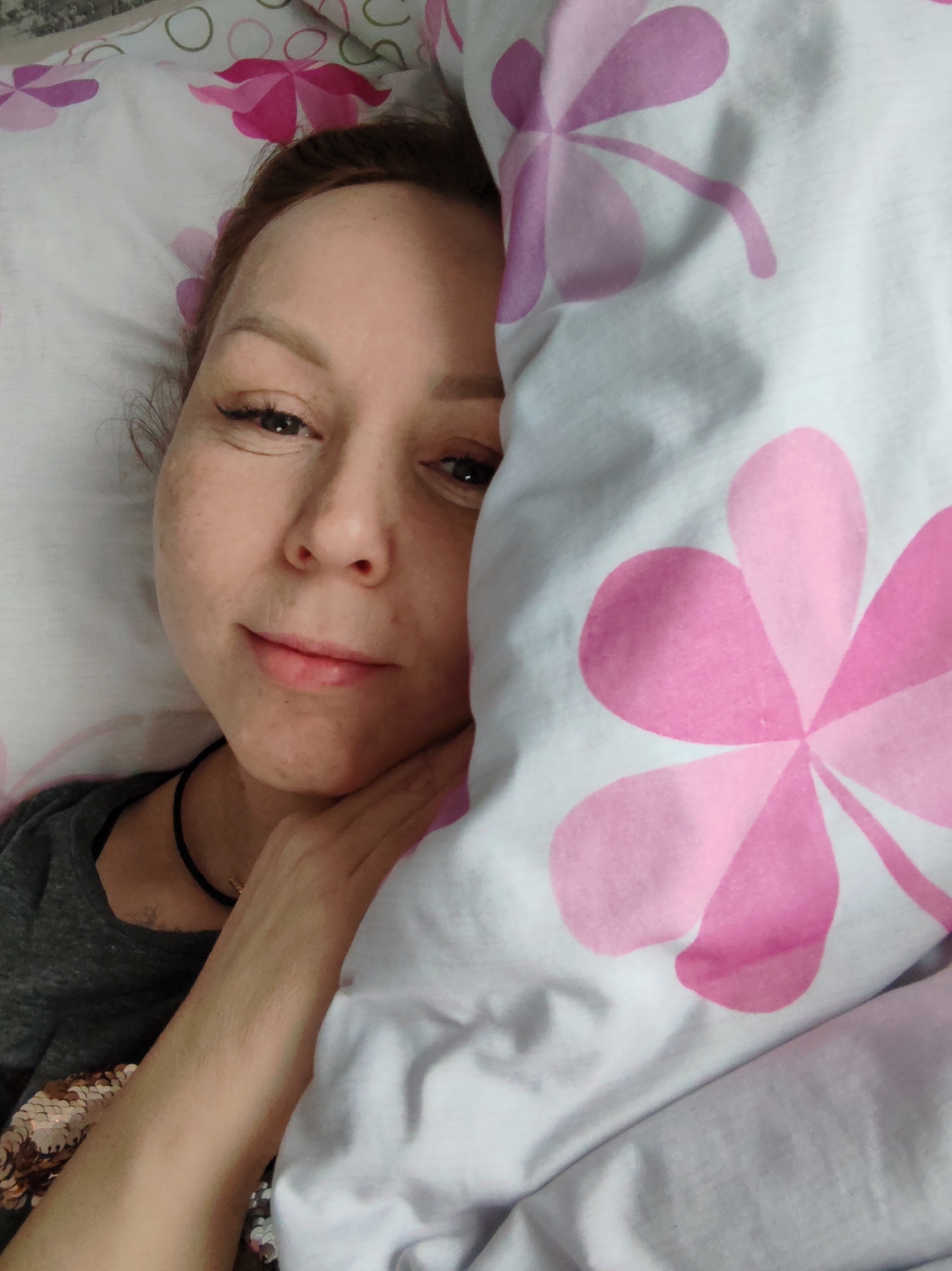 A person lies in bed, with a floral-patterned pillow next to their face. The person is fair-skinned with brown hair, and their hand is resting on their cheek. They are wearing a gray shirt with gold sequins on the shoulder area. The bedding has a pattern of pink four-petal flowers on a white background. The location is potentially Chaikovsky, Russia.