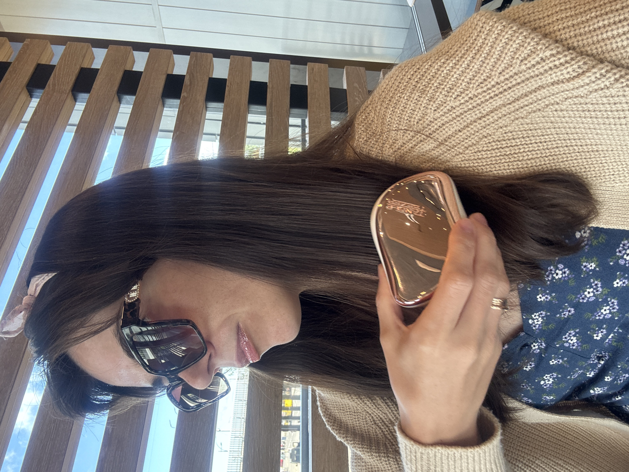 In this medium shot image, a woman with dark hair and a headband sits indoors, looking to her left and off-camera. She is wearing large, dark sunglasses with a leopard print frame and a light brown, knitted cardigan over a dark blue floral-print dress. Her right hand is raised, holding a small, rose-gold, handheld mirror. A ring is visible on her left ring finger. The background features a wall made of horizontal wooden slats with vertical black supports, behind which a bright sky and some structures are visible through the gaps. The lighting suggests it is daytime.