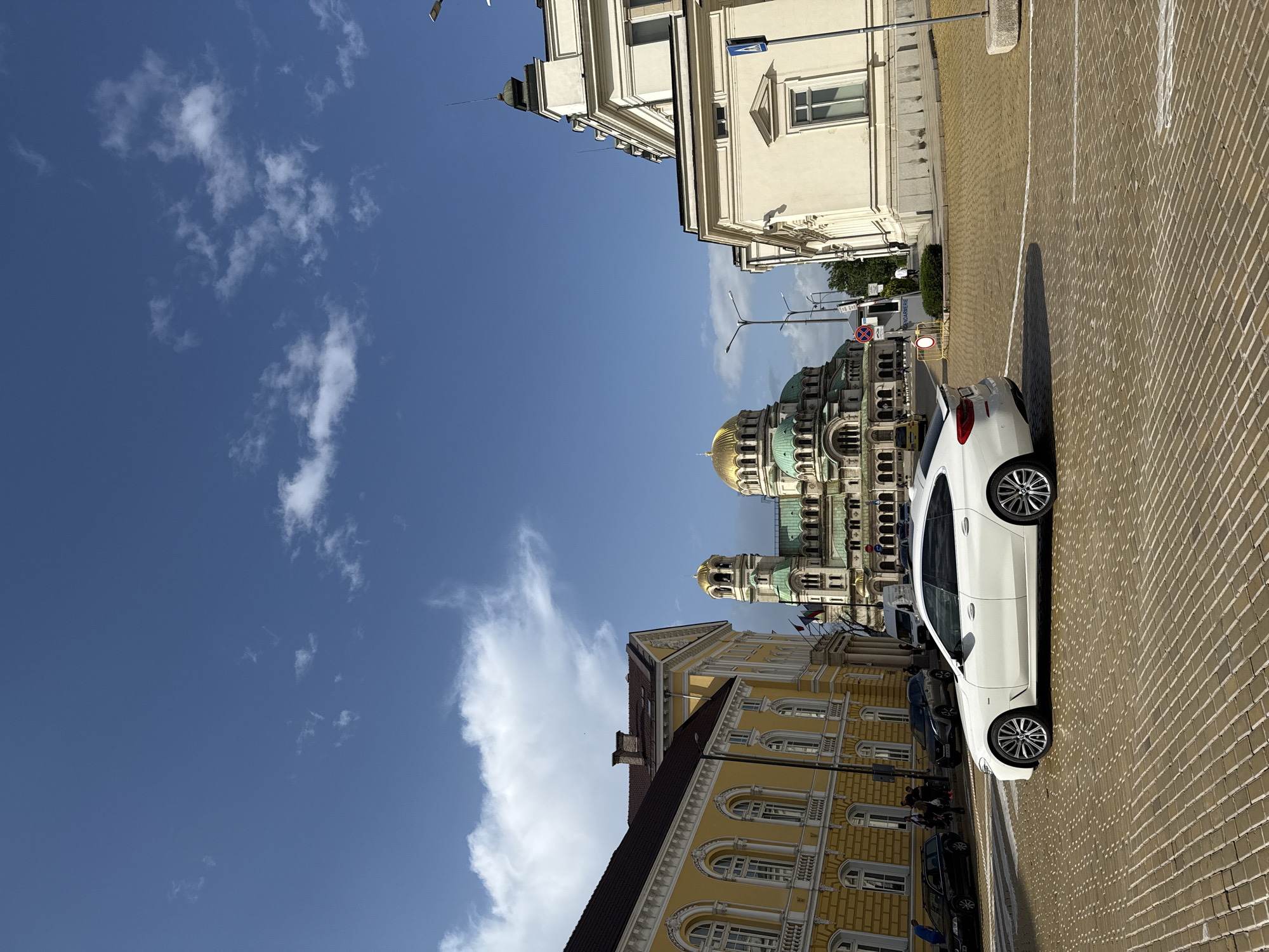 The image depicts an urban scene in Sofia, Bulgaria, dominated by architectural landmarks under a partly cloudy sky. A white car is parked on a cobblestone street, oriented vertically in the frame. In the background, the Alexander Nevsky Cathedral is visible with its prominent golden domes and detailed façade. Adjacent to the cathedral is a yellow building, possibly a governmental structure, featuring multiple arched windows and ornate detailing. On the other side of the car, part of another building is visible. The sky is bright blue with scattered white clouds.