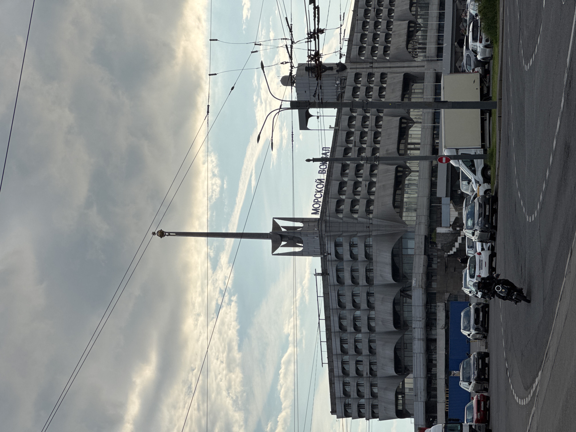 The image displays an urban landscape featuring a large, grey, brutalist-style building, which is prominently identified by the Cyrillic text "МОРСКОЙ ВОКЗАЛ" (Morskoy Vokzal) on its facade, translating to "Marine Terminal" or "Sea Station." This structure, located in Saint Petersburg, Russia, exhibits a repetitive design with numerous curved window alcoves and is topped with a tall, slender, mast-like element.
The sky above is partially cloudy, showing a mix of grey clouds and patches of brighter blue, indicative of daytime. A network of power lines and trolley wires intersects the upper portion of the frame. In the foreground, a wide road or parking area runs parallel to the building. This area is lined with various parked vehicles, including several cars, vans, and a large white truck with a beige cargo unit. A red circular traffic sign is also visible among the parked vehicles. On the right side of the road, a person is seen riding a dark-colored motorcycle. The overall scene captures an ordinary moment in an urban environment, with no particular activity or strong emotions conveyed.
