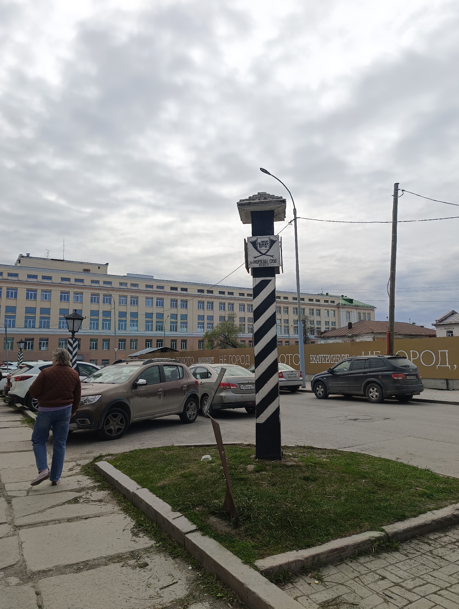 The image depicts a street scene in Yekaterinburg, Russia, characterized by overcast skies and a mix of architectural styles. In the foreground, a pedestrian is walking on a paved sidewalk. Several cars are parked along the street, including a brown SUV and a black SUV.
A notable feature is a striped black and white post or monument with a sign attached; the sign displays the inscription "ДО МОСКВЫ 1766 ВЕРСТ." A streetlight is positioned near the post. The background features a large, multi-story building with numerous windows. Other structures and power lines are also visible. The overall environment suggests an urban setting with a blend of historical and modern elements.