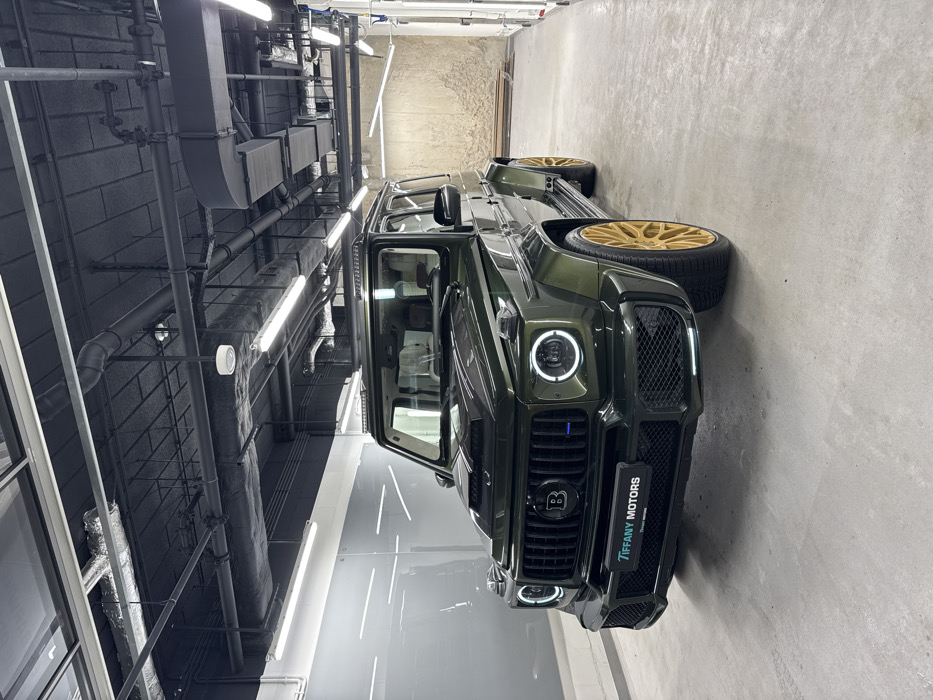The image displays a dark olive green modified Mercedes-Benz G-Wagen, identified by Brabus badging on the front grille, parked centrally within an indoor facility in Warsaw, Poland. The vehicle features circular LED daytime running lights, a prominent hood scoop, a custom front bumper with mesh grilles, and multi-spoke gold-colored wheels. A "TIFFANY MOTORS" plate frame is visible on the lower front bumper.
The environment consists of a light grey concrete floor. The wall to the left is composed of reflective, light grey panels, while the wall to the right is textured, light brown/beige, resembling stucco or rough concrete, with several light-colored wooden planks leaning against it in the background. The ceiling is painted black, exposing extensive industrial ductwork, electrical conduits, and numerous linear fluorescent light fixtures, along with some circular ceiling lights, providing bright, artificial illumination. No individuals are present, and no actions or interactions are depicted in the static scene.
