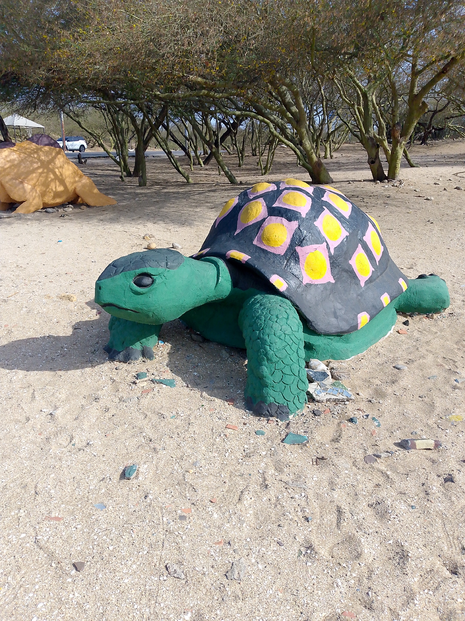 The image captures a life-sized, painted concrete sculpture of a turtle in a sandy area. The turtle's body is green, while its shell is black with pink and yellow markings. The turtle is located in Coro, Venezuela, near other turtle sculptures and trees. A white car and a tent are visible in the background. The ground is a mixture of sand and debris.