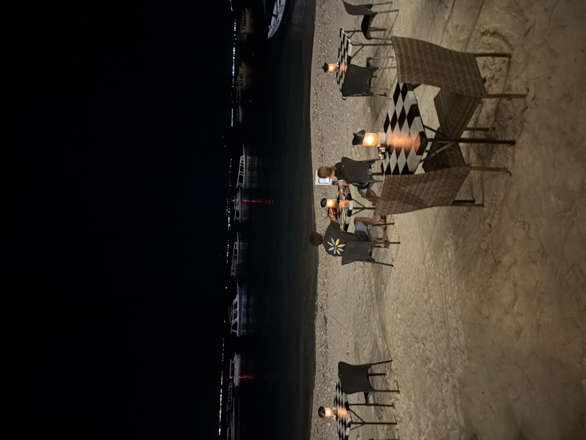 An outdoor scene at night features a beachside dining area with several tables and chairs set up on the sand. The ocean is visible in the background with numerous boats anchored offshore. Two individuals are seated at a table in the middle ground, facing each other, with one appearing to use a laptop. Two other tables are also set with checkered tablecloths and lanterns, one on the left and one on the right. The beach is lit by the lanterns and ambient light from the boats. The location is the island of Gili Air, Indonesia.