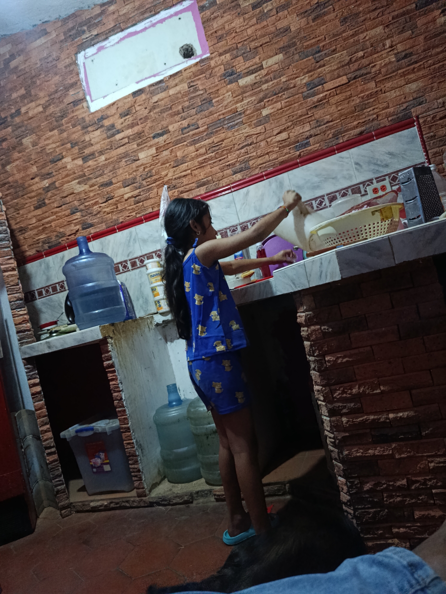 A young girl wearing a blue pajama set with a teddy bear print stands in a kitchen in Juan Griego, Venezuela. The kitchen has a brick-patterned wall and a countertop with a sink, dish rack, and various bottles and containers. A large blue water jug is visible on a shelf below the counter. The girl is holding a white object, possibly dough, over a yellow basket. The floor is tiled. The lighting is dim, suggesting it might be evening or the room is not well-lit.